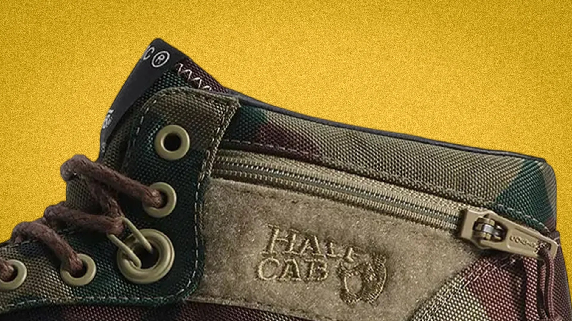 Close-up of a camouflage fabric shoe with brown laces, metal eyelets, and a suede patch with "HALF CAB" embossed next to a zipper.