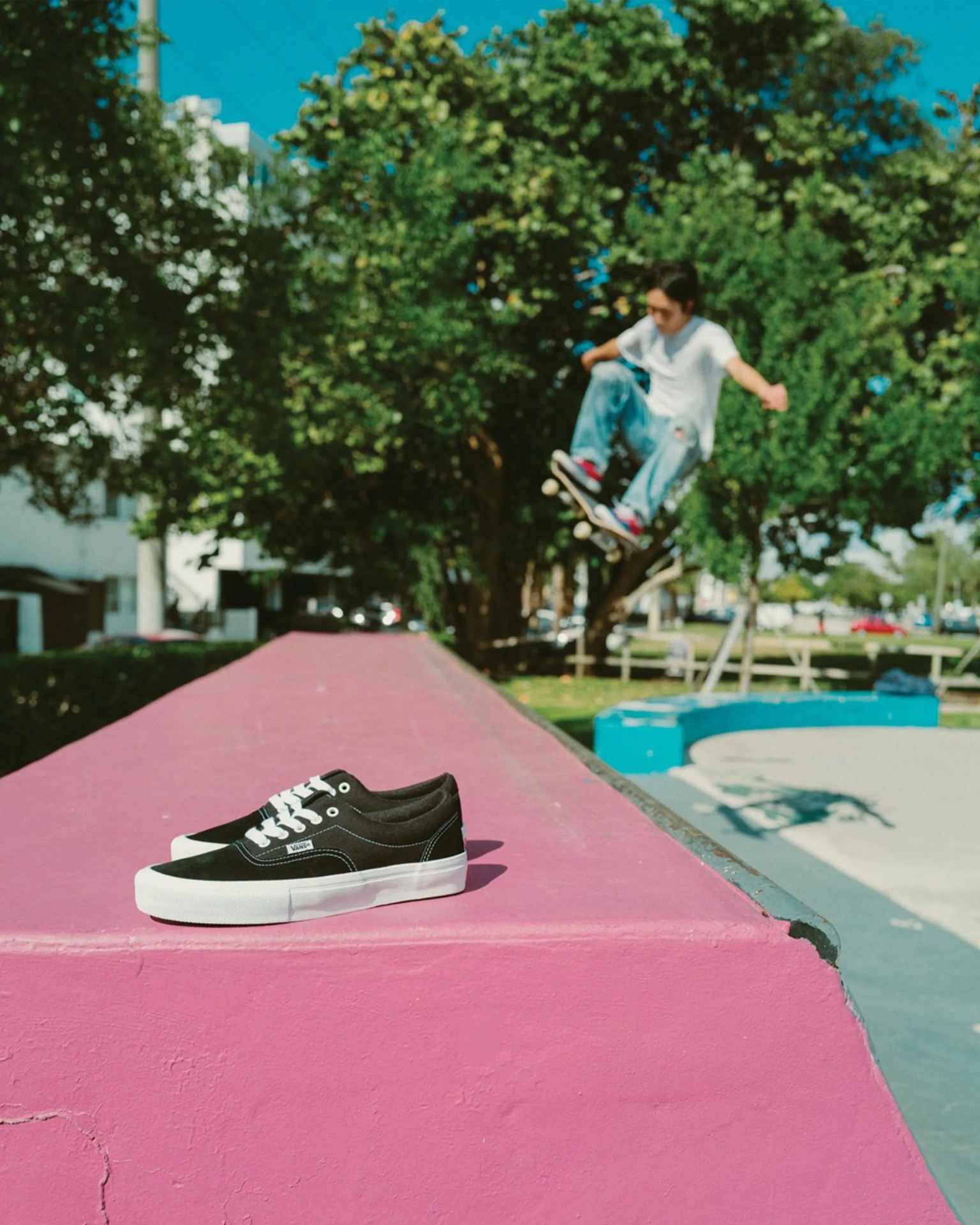 Black and white Vans sneakers on a pink ledge with a skateboarder in mid-air in the background.