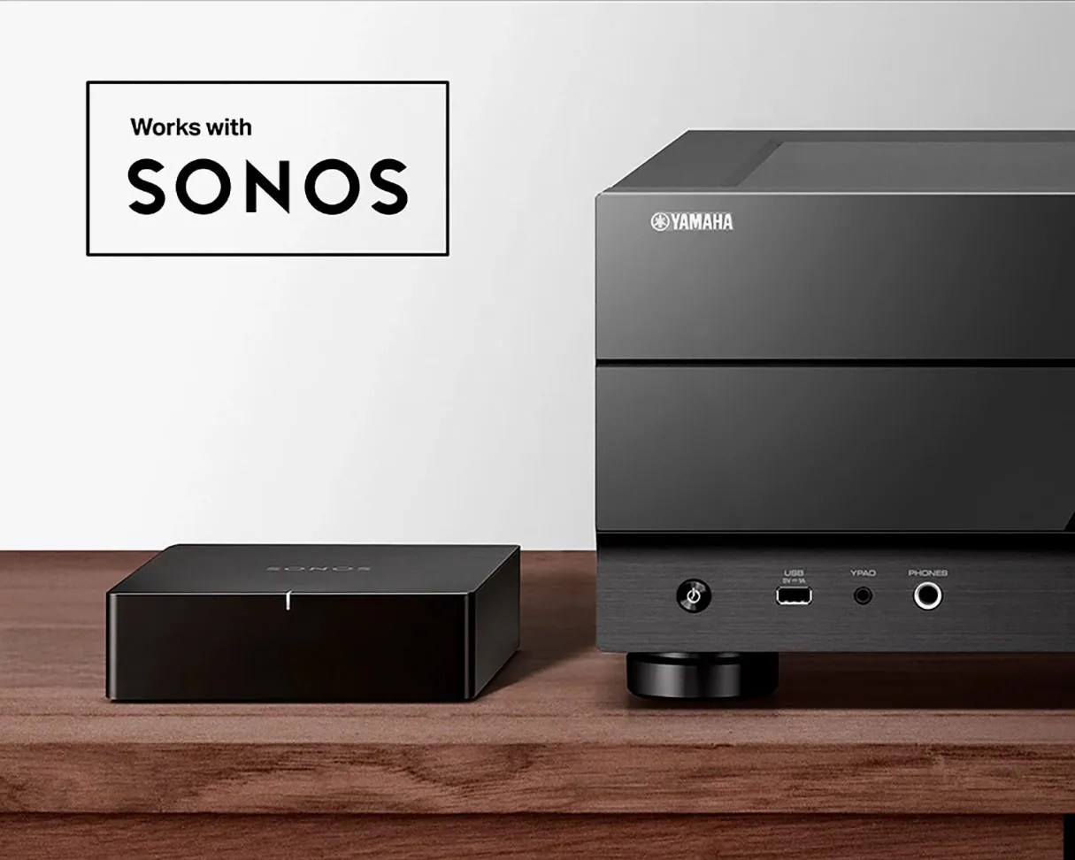Black Sonos device and black Yamaha audio equipment on a wooden surface with "Works with Sonos" text.