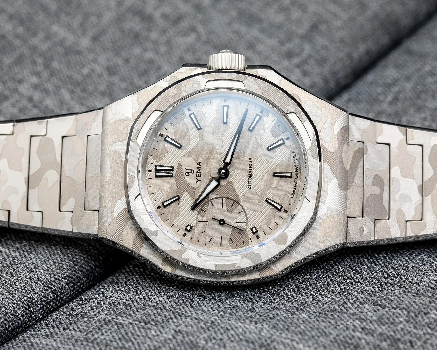 Silver camouflage-patterned Yema automatic wristwatch with a matching metal bracelet on gray fabric.