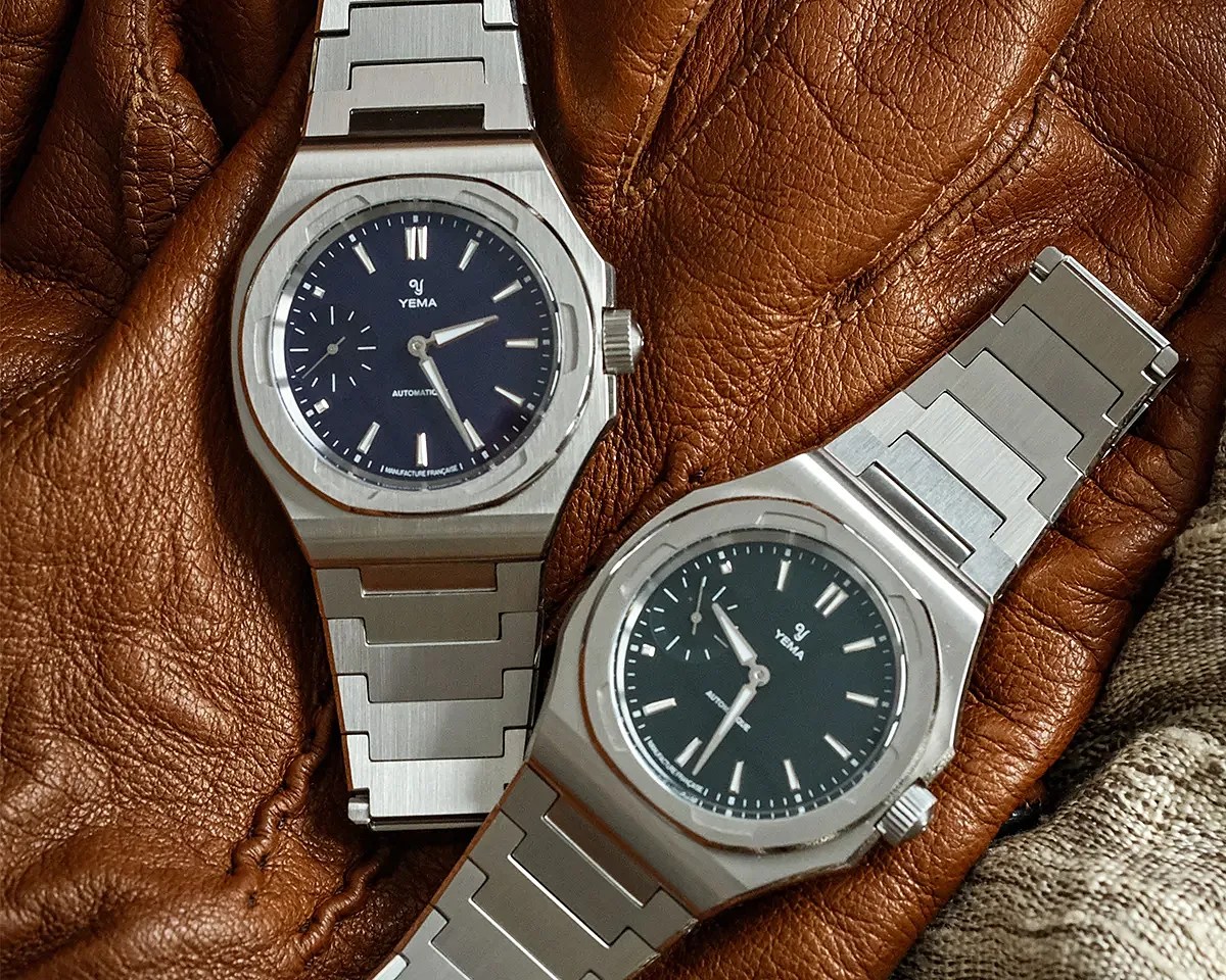Two silver Yema automatic wristwatches with black dials and stainless steel bracelets on a brown leather surface.