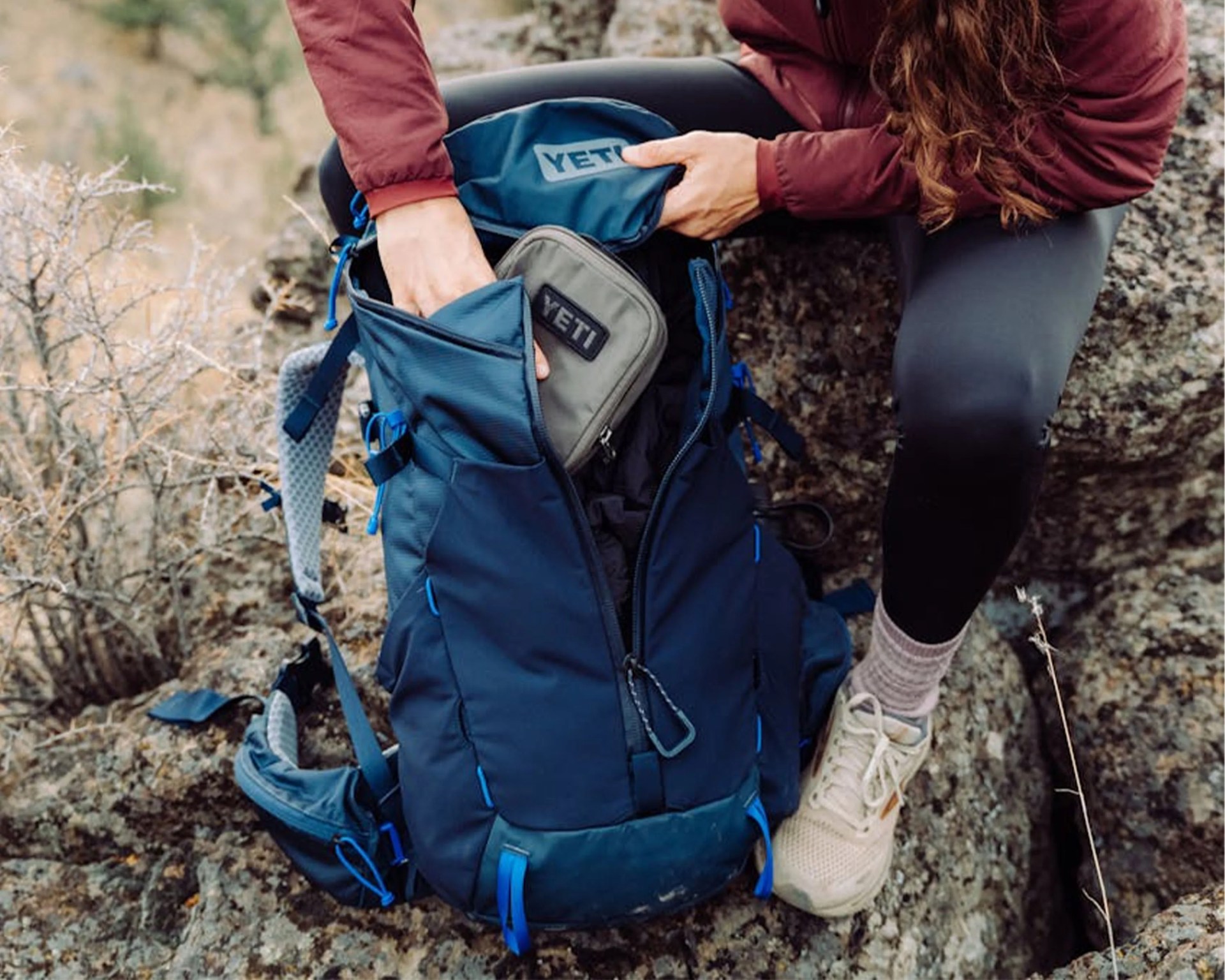Blue YETI backpack with multiple compartments being opened by a person sitting on rocky terrain.