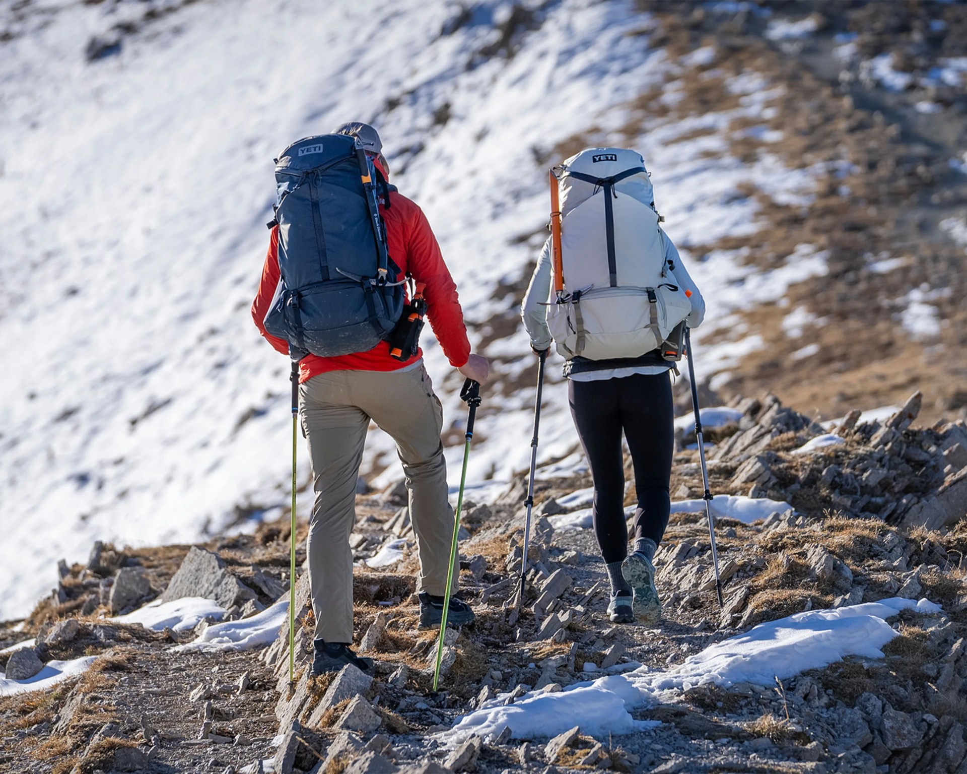 Two hikers with large YETI backpacks and trekking poles walking on a rocky, snowy mountain trail.