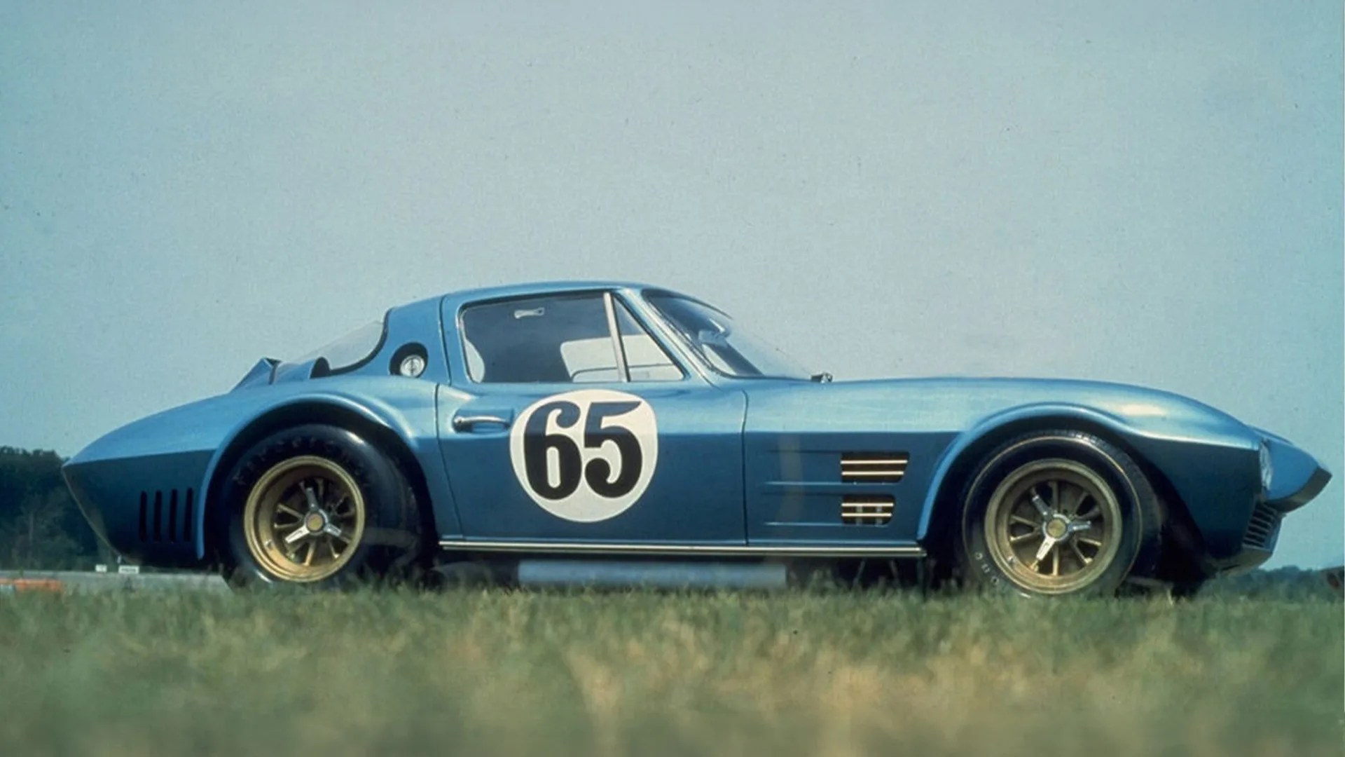 Blue vintage race car with number 65 on the door and gold wheels parked on grass.