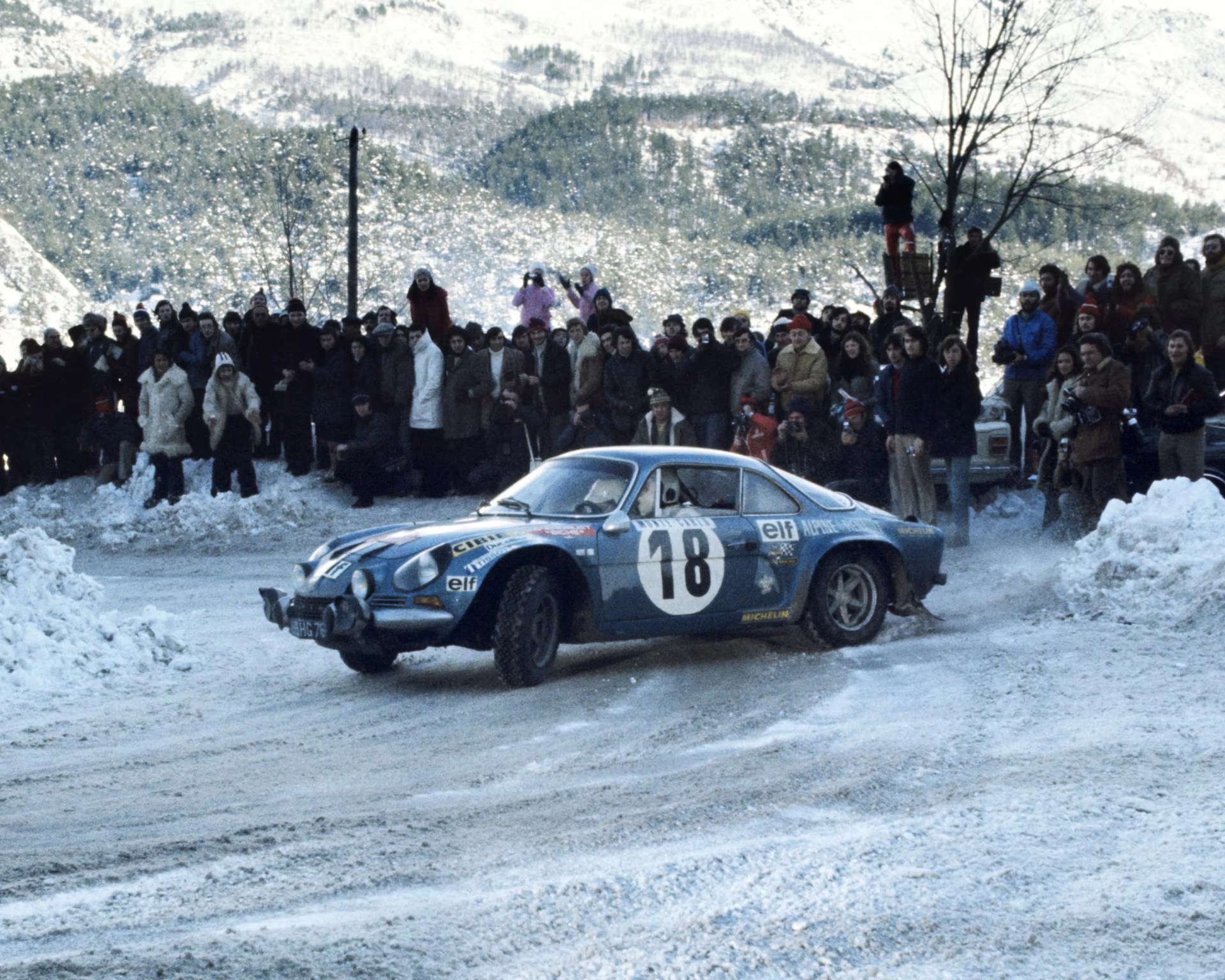 Blue Alpine A110 rally car with number 18 drifting on a snowy road surrounded by spectators.
