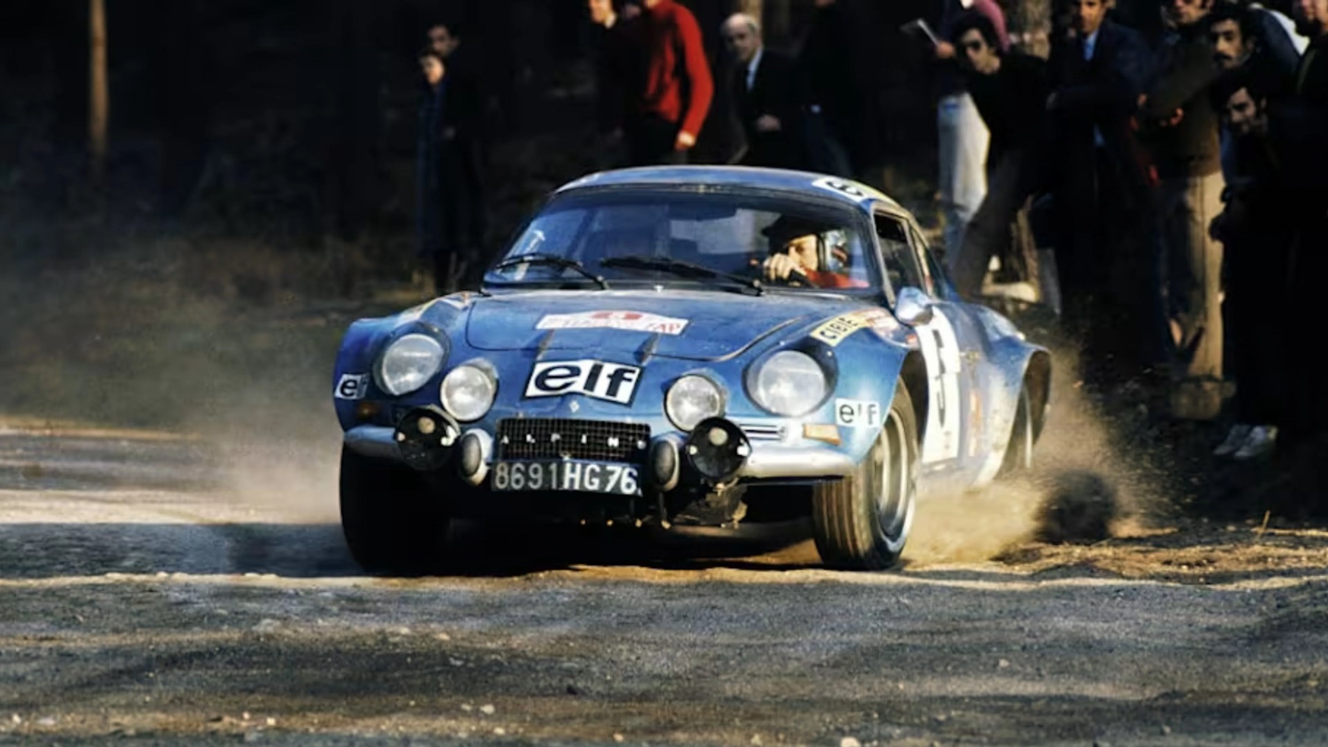 Blue Alpine A110 rally car with multiple round headlights kicking up dust on a gravel road.