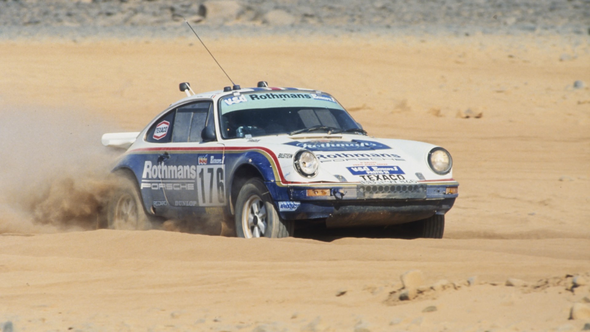 White and blue Rothmans Porsche rally car number 176 kicking up sand in a desert setting.