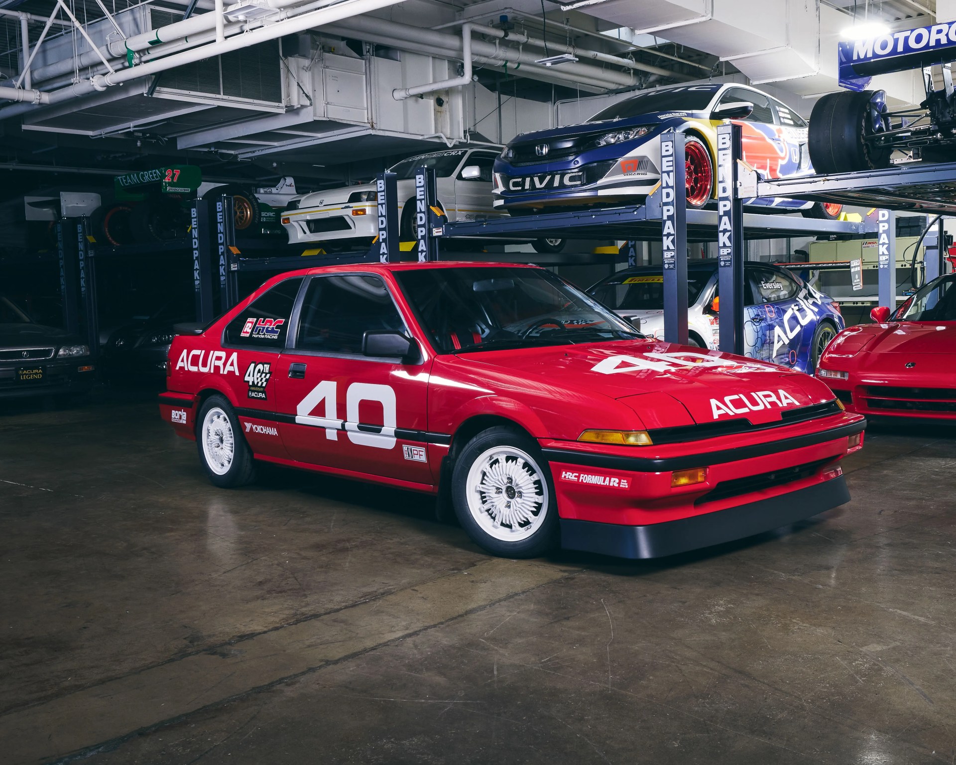 Red Acura race car with white number 40 and white wheels in a garage with other cars on lifts.