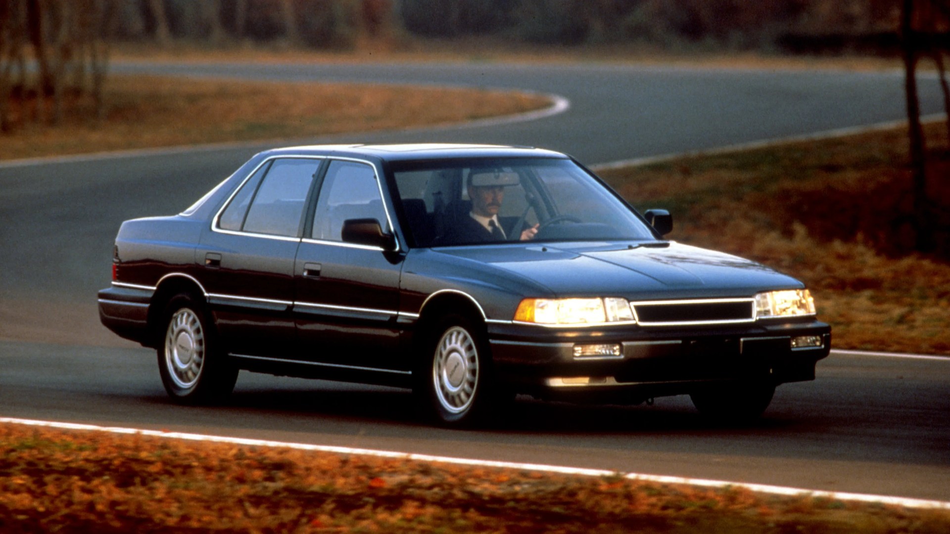 Dark blue four-door sedan driving on a curved road with headlights on and a person inside.