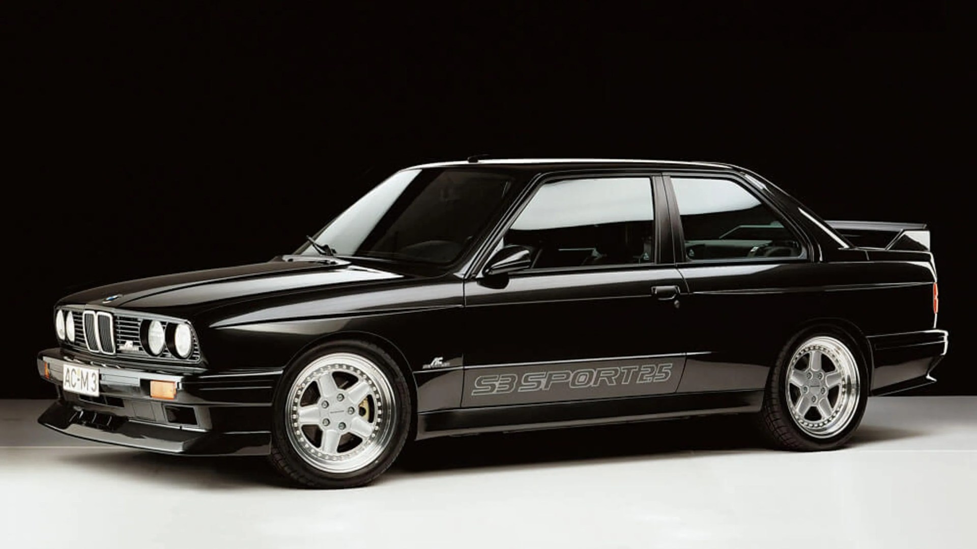 Glossy black BMW E30 M3 with silver five-spoke wheels and "S3 SPORT25" decal on the side.