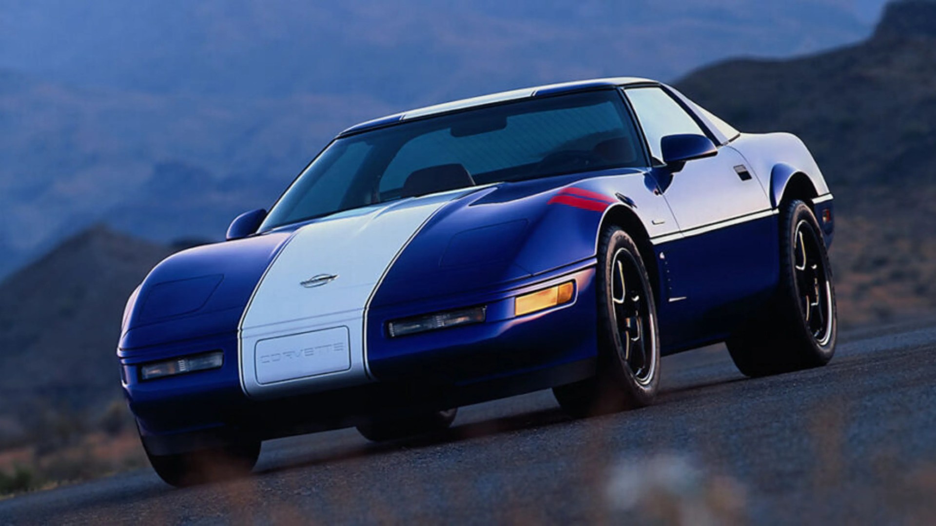 Blue and white Chevrolet Corvette sports car with red stripes on front fender parked on a road.