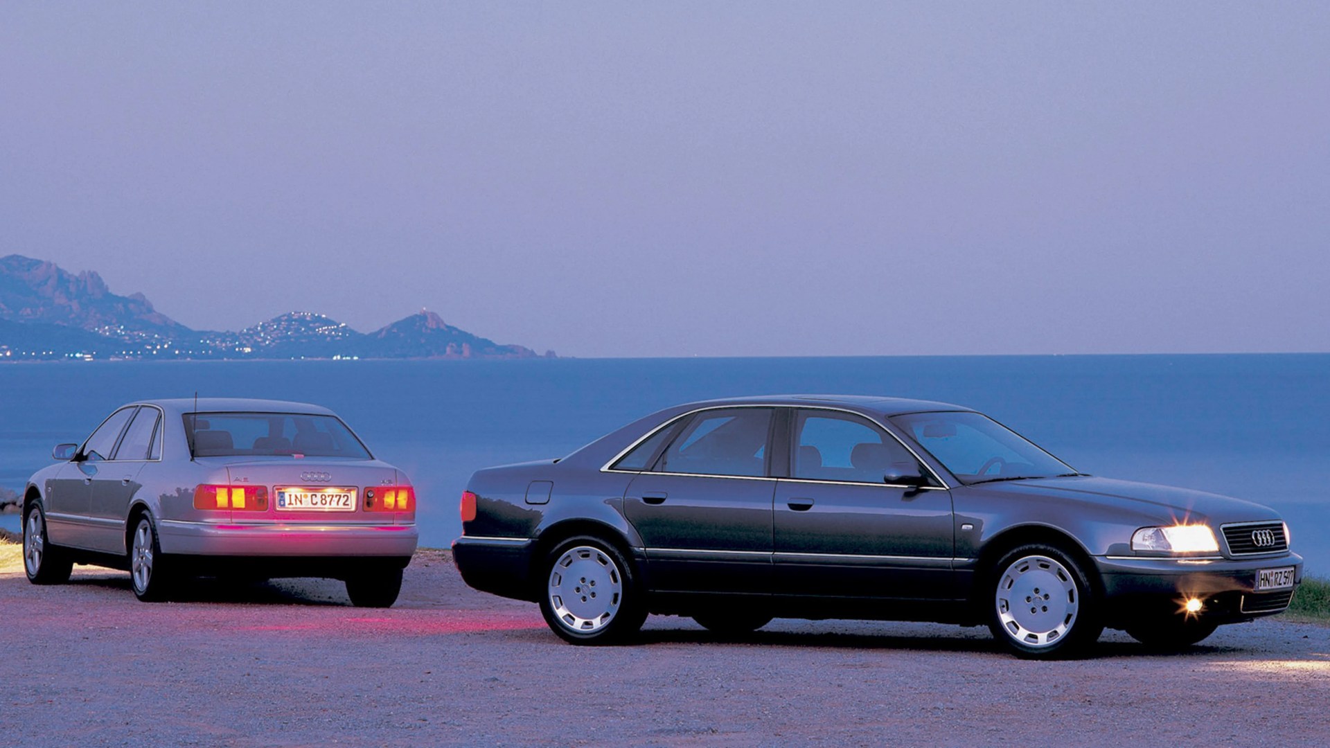 Two Audi A8 sedans, one silver rear view with taillights on and one black side view with headlights on, parked near a body of water at dusk.