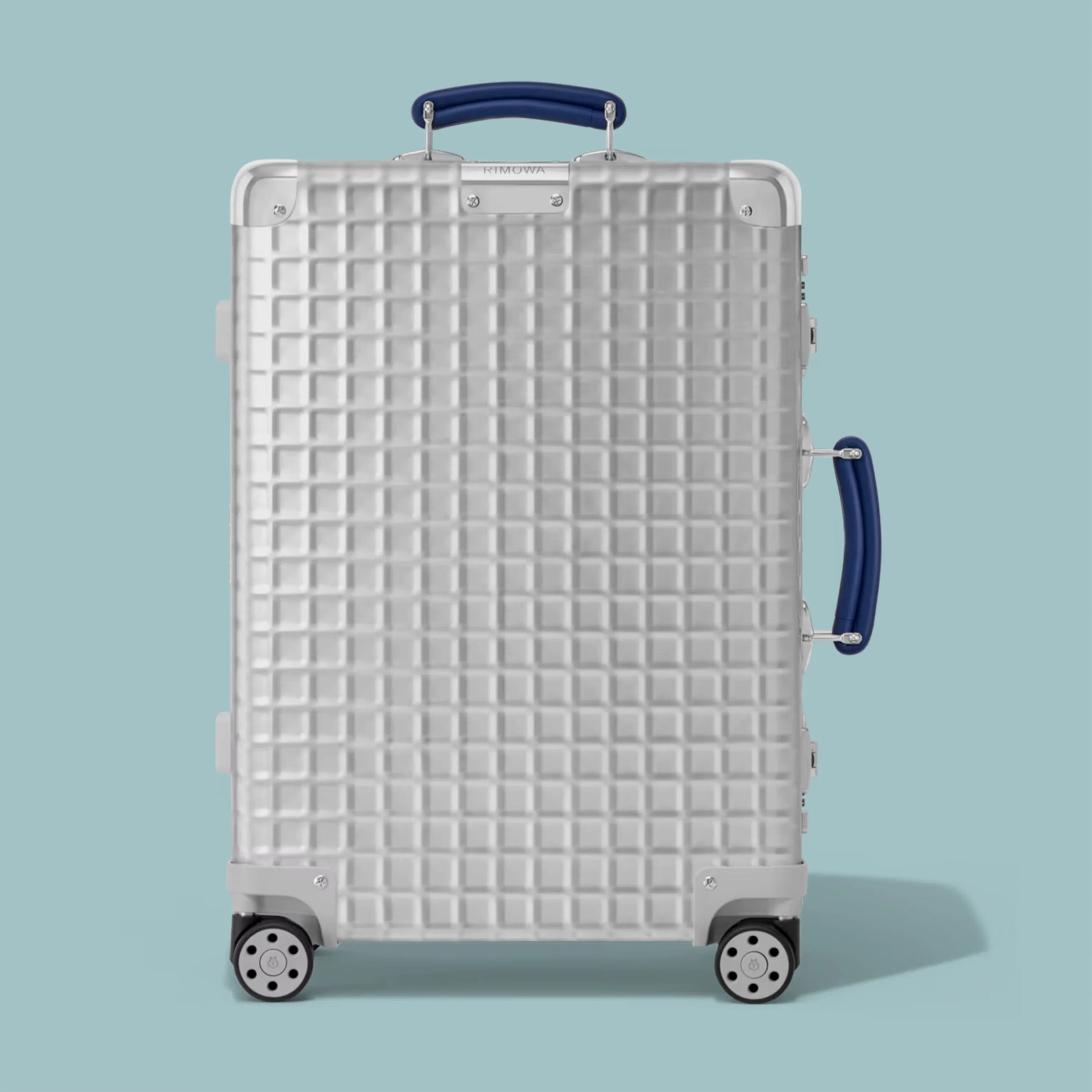 Silver hard-shell suitcase with a textured grid pattern, blue handles, and four wheels on a light blue background.