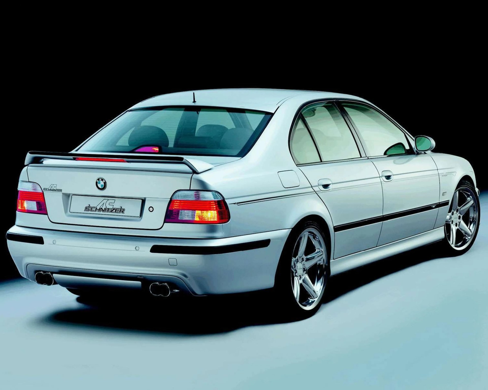 Silver BMW sedan with AC Schnitzer tuning, rear spoiler, and chrome five-spoke wheels on a light surface.