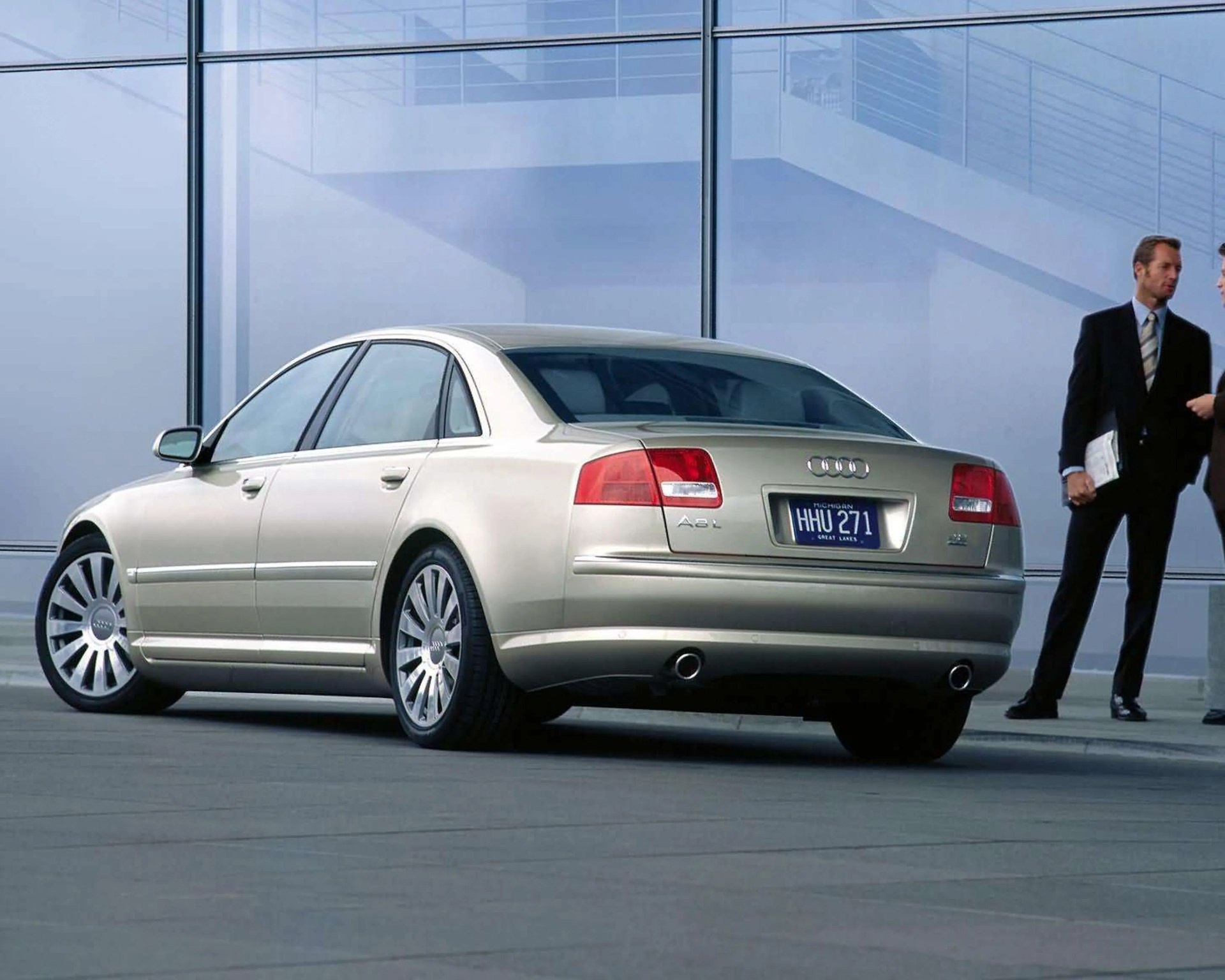 Silver Audi A8L sedan with dual exhausts parked indoors near glass walls and two men in suits standing nearby.