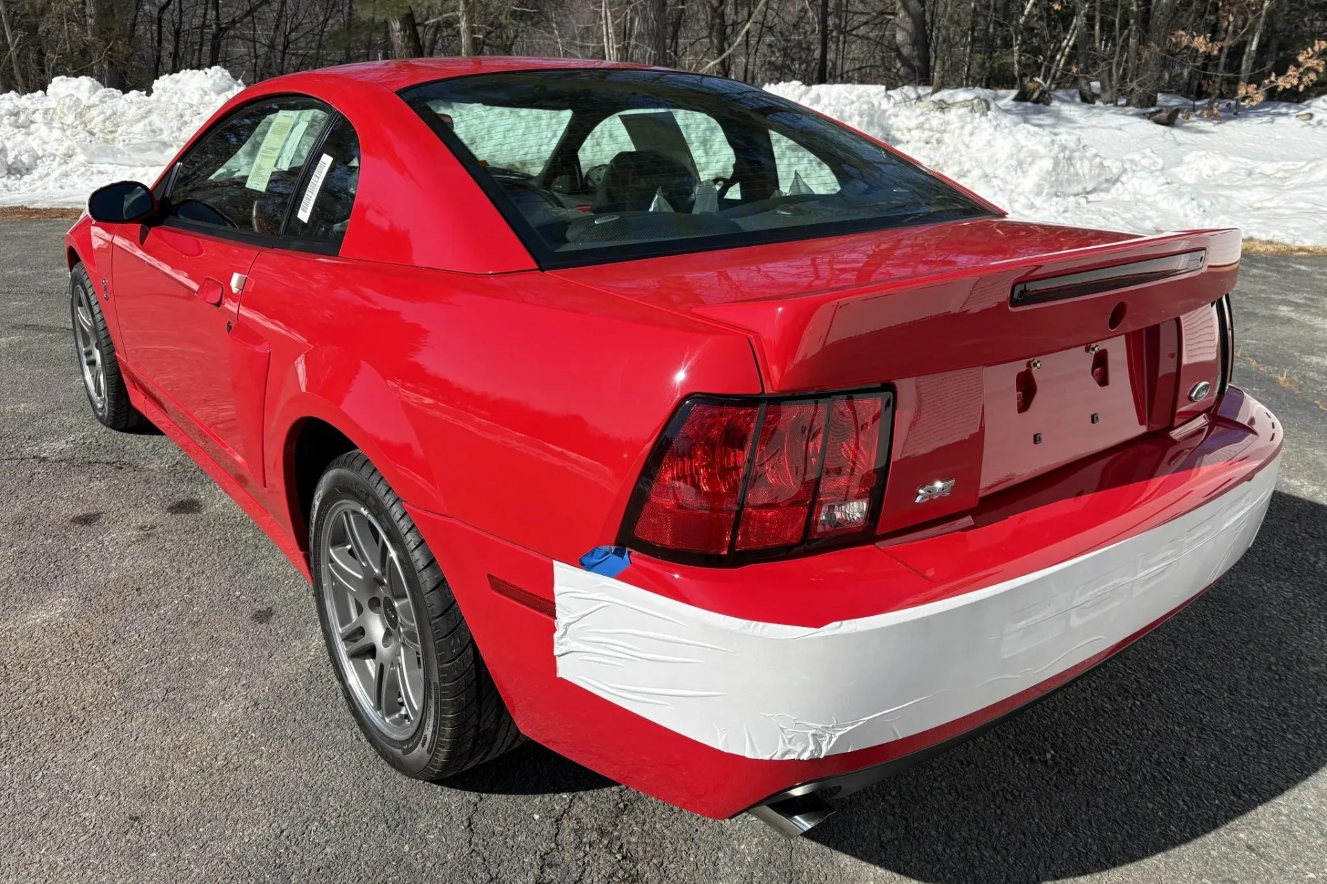 Red Ford Mustang SVT Cobra with white protective tape on rear bumper parked on asphalt with snow in background.