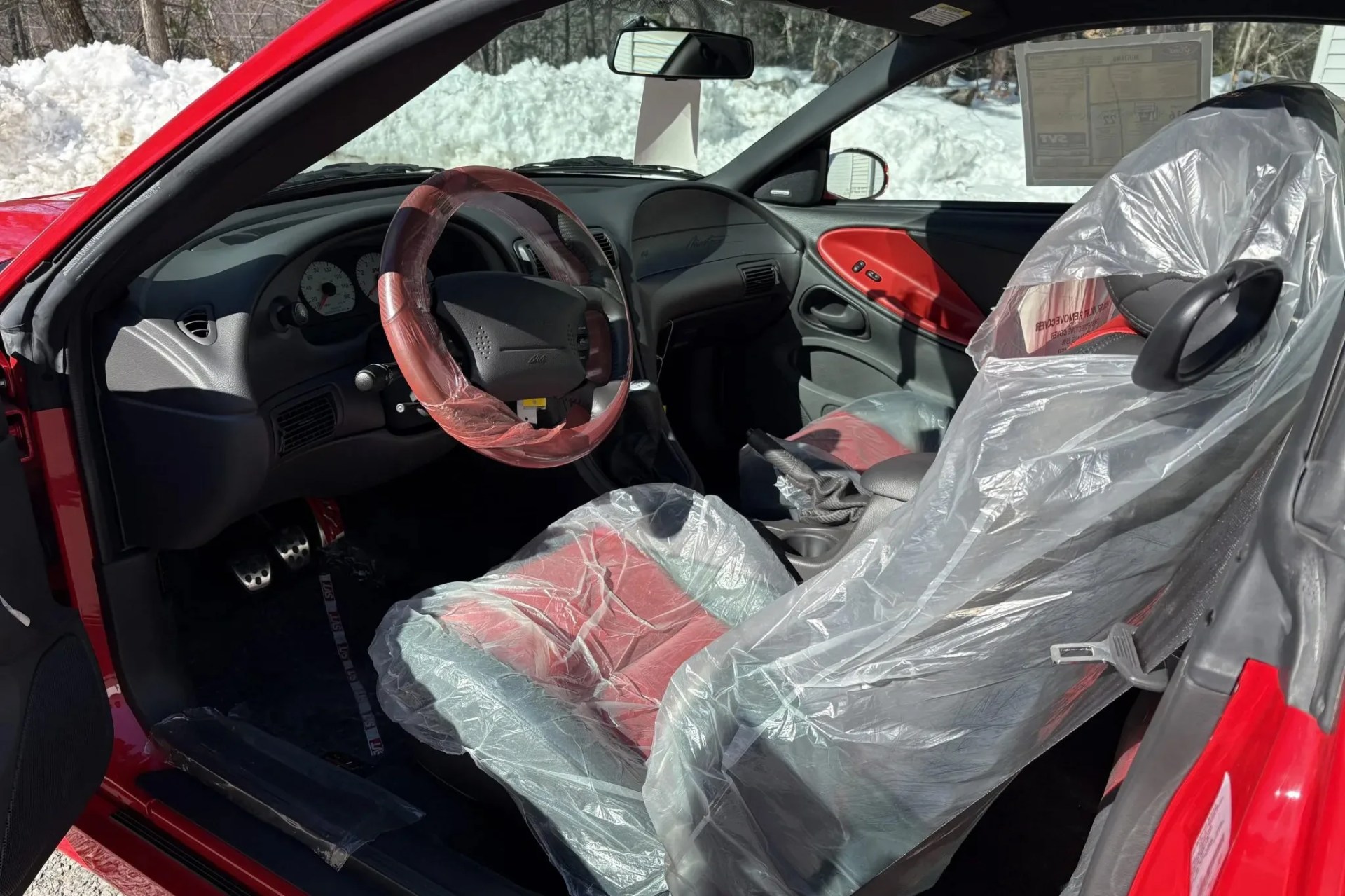 Red and black car interior with plastic covers on the seats and steering wheel, and snow visible outside.