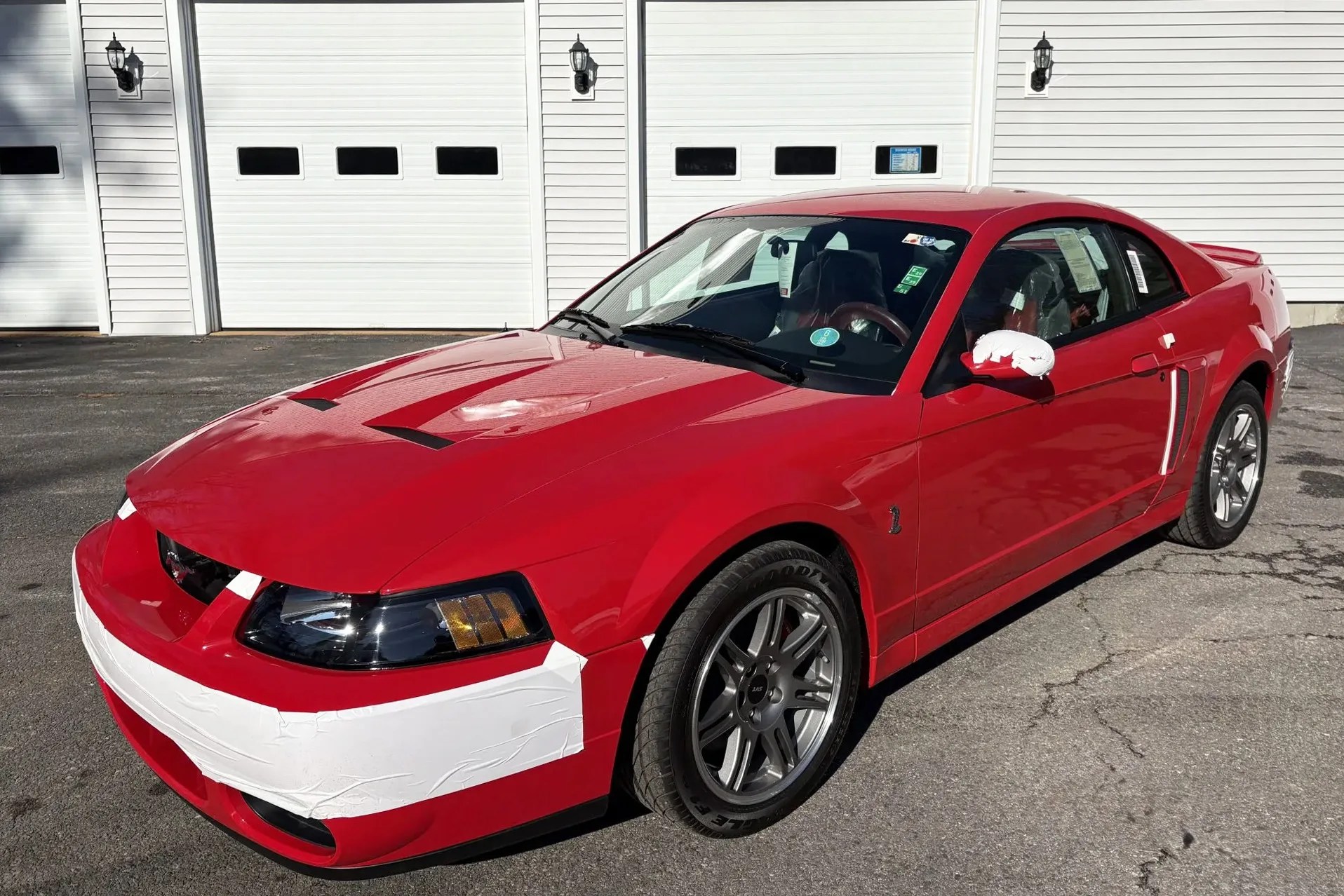 Red Ford Mustang Cobra with white protective tape on front bumper and side mirror, parked on asphalt.