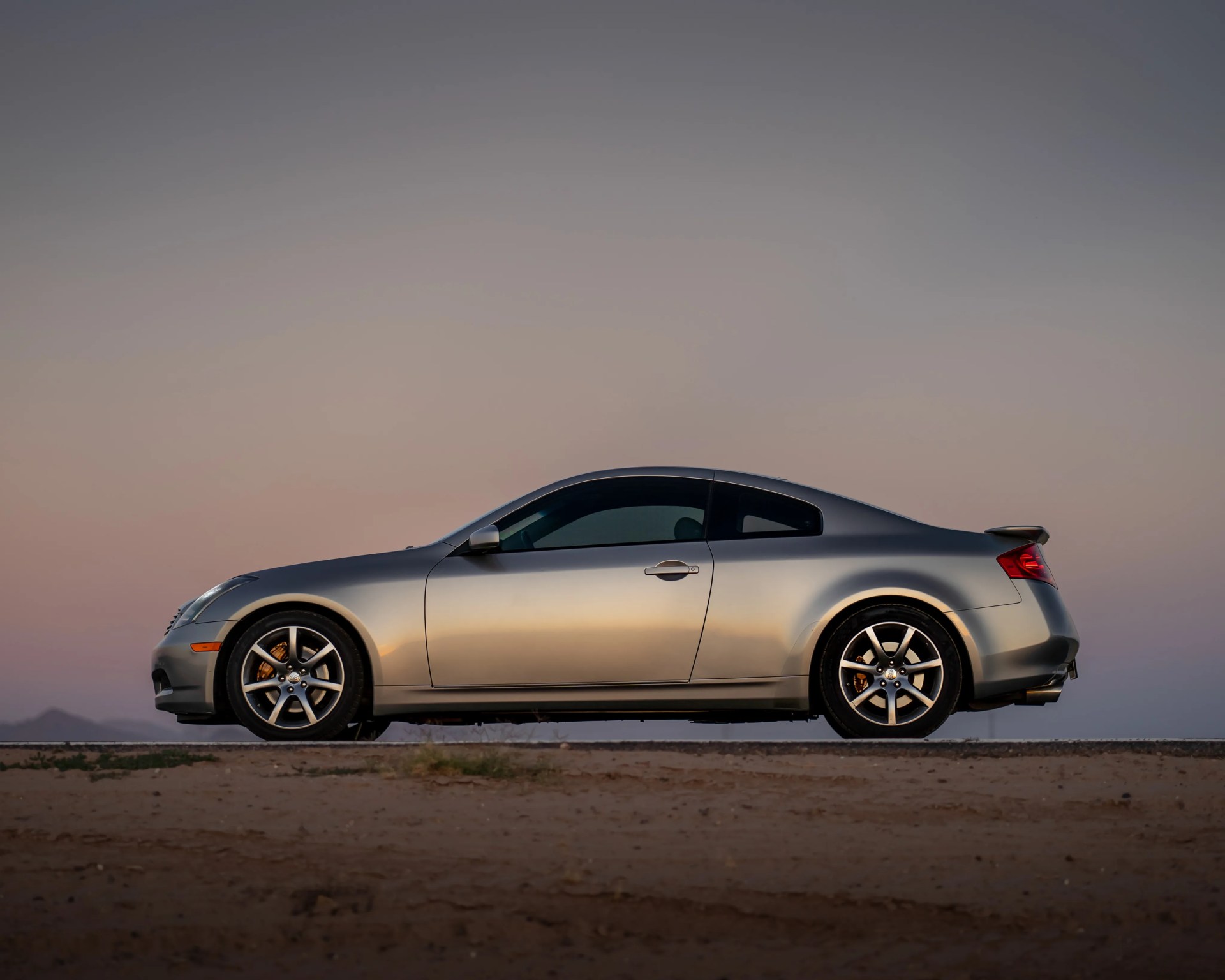 Silver two-door sports coupe parked on a desert road at sunset with tinted windows and alloy wheels.