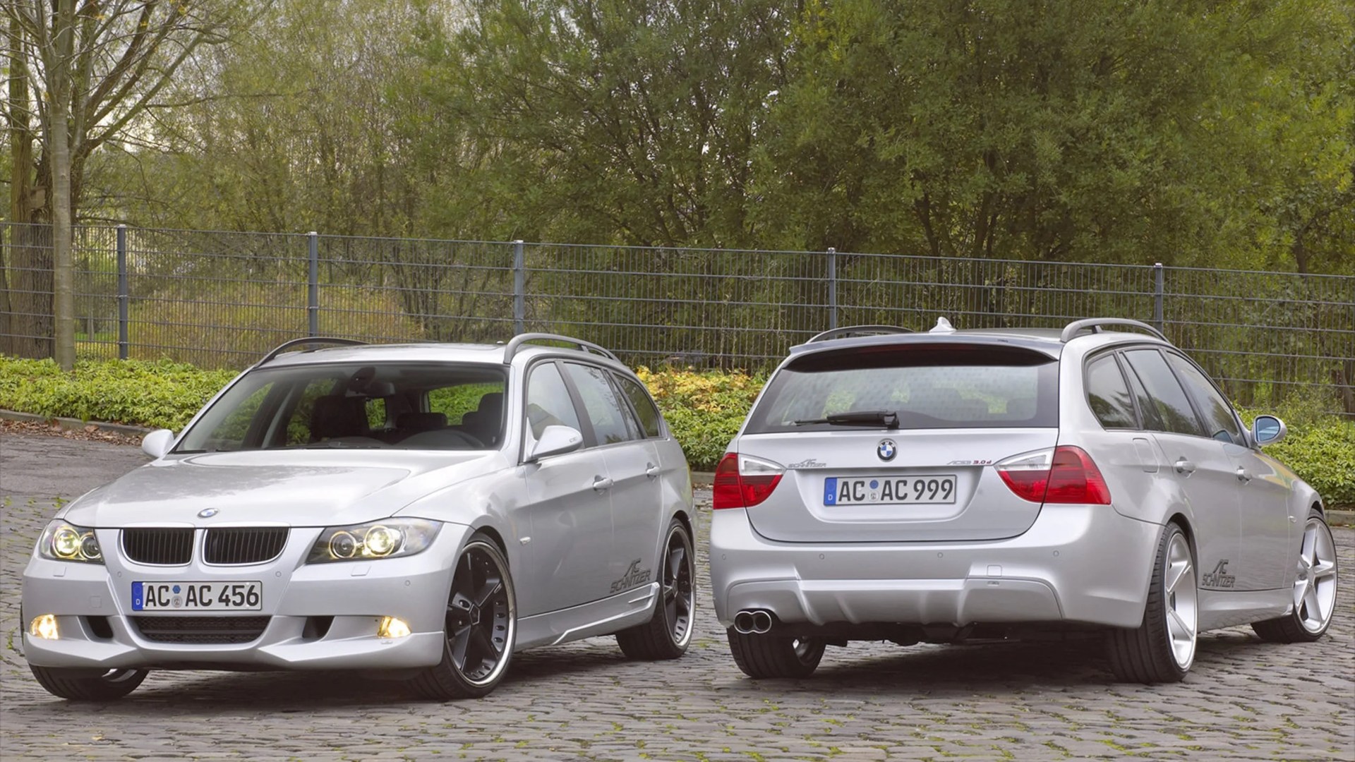 Two silver BMW station wagons with AC Schnitzer tuning parked on cobblestone pavement in front of a metal fence and trees.