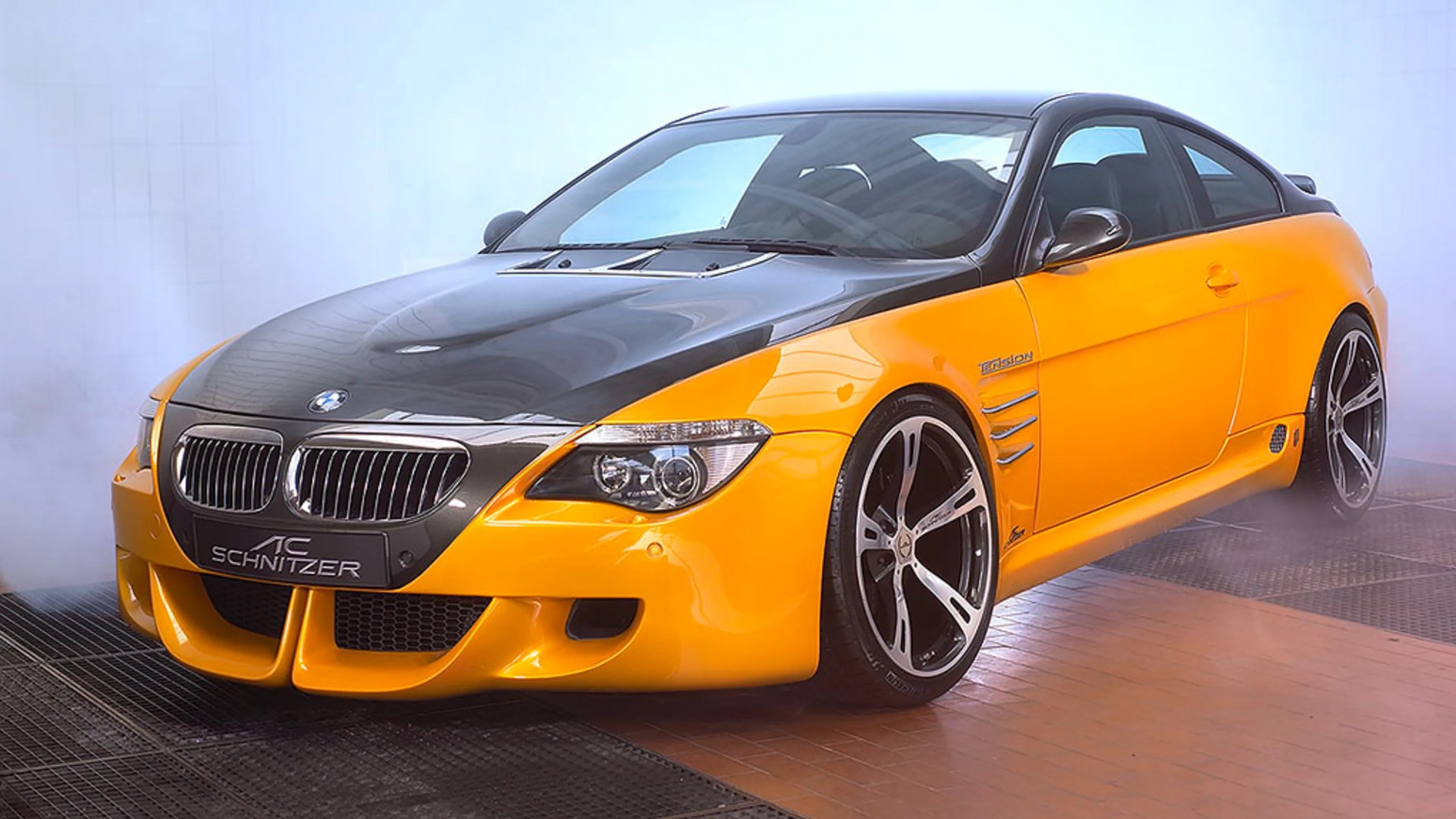 Yellow and black BMW coupe with AC Schnitzer tuning and large black and silver wheels in a smoky garage.