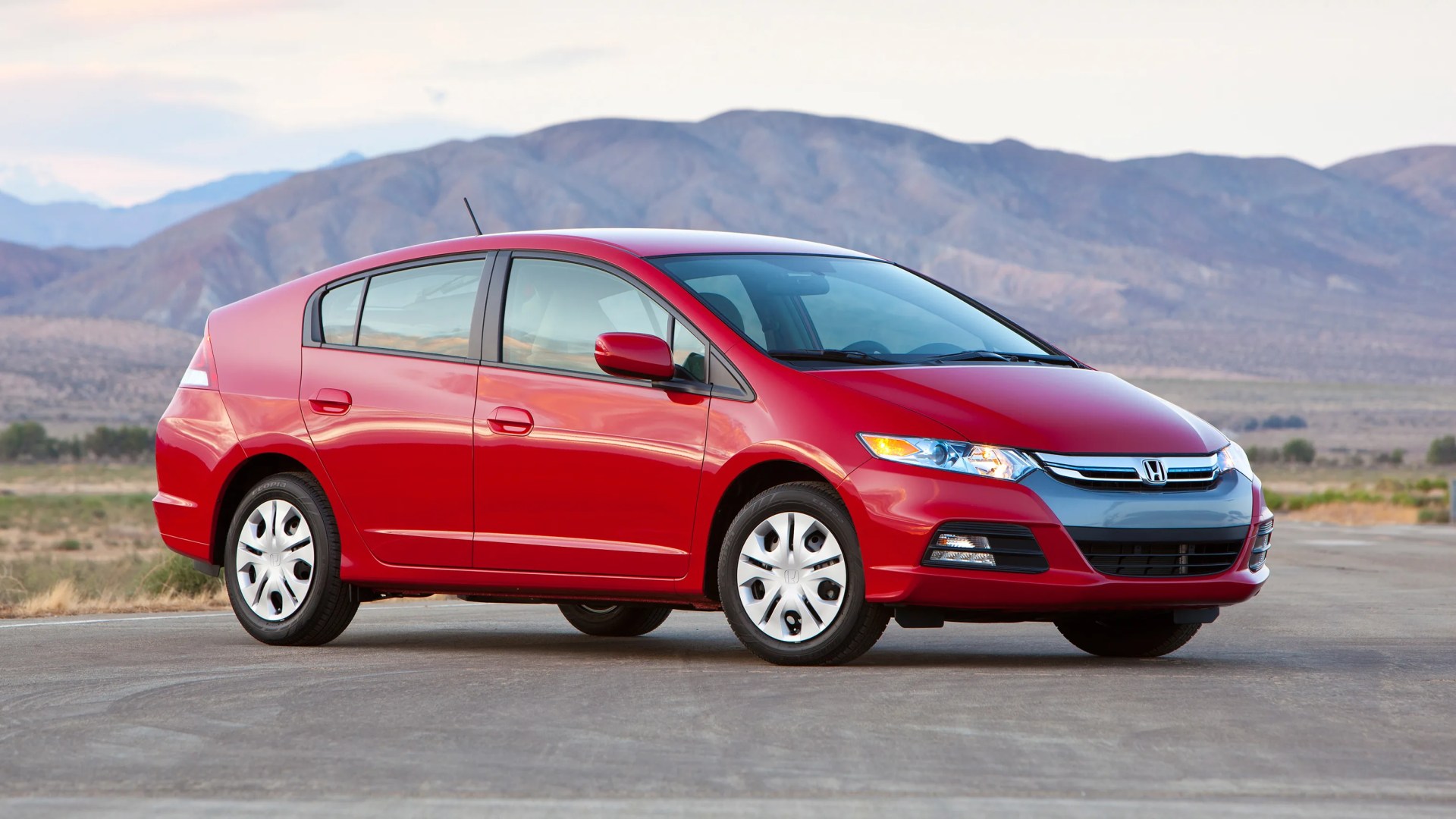 Red Honda Insight hybrid hatchback parked on a road with mountains in the background.