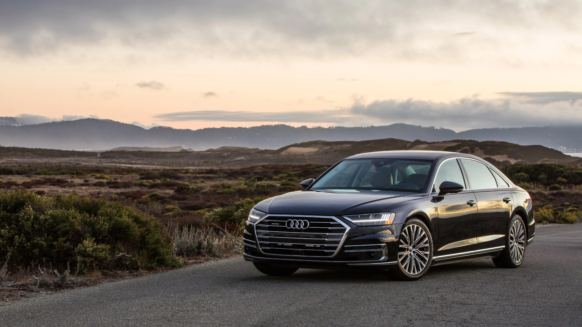 Black Audi sedan parked on a road with hills and cloudy sky in the background at sunset.