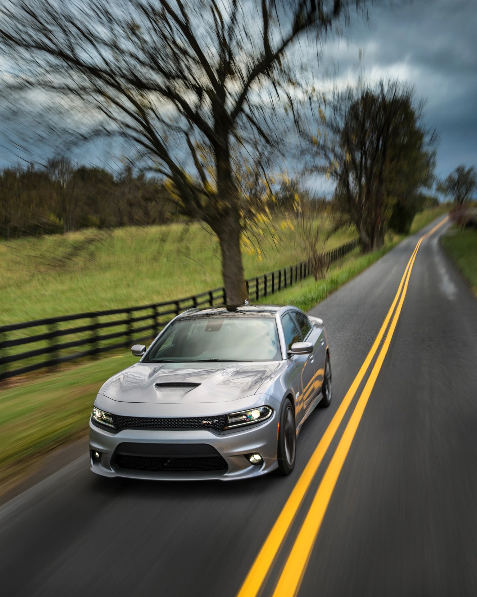 Silver Dodge Charger SRT driving on a rural road with yellow double lines and blurred trees in the background.