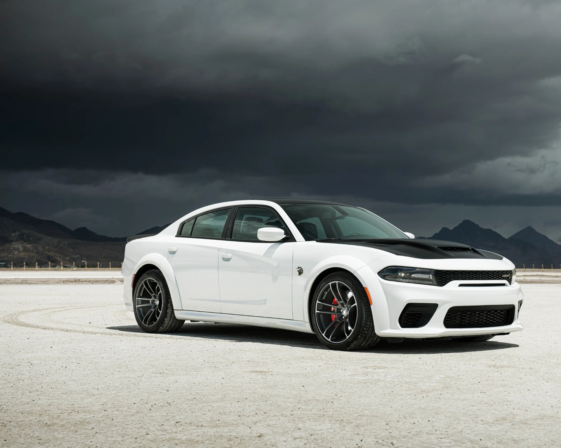 White Dodge Charger Hellcat with black hood and red brake calipers on a salt flat under dark cloudy sky.