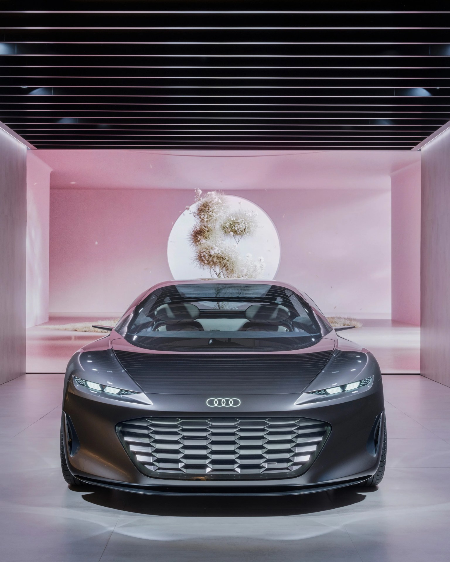 Matte gray Audi concept car with illuminated logo and large grille in a modern showroom with pink walls.