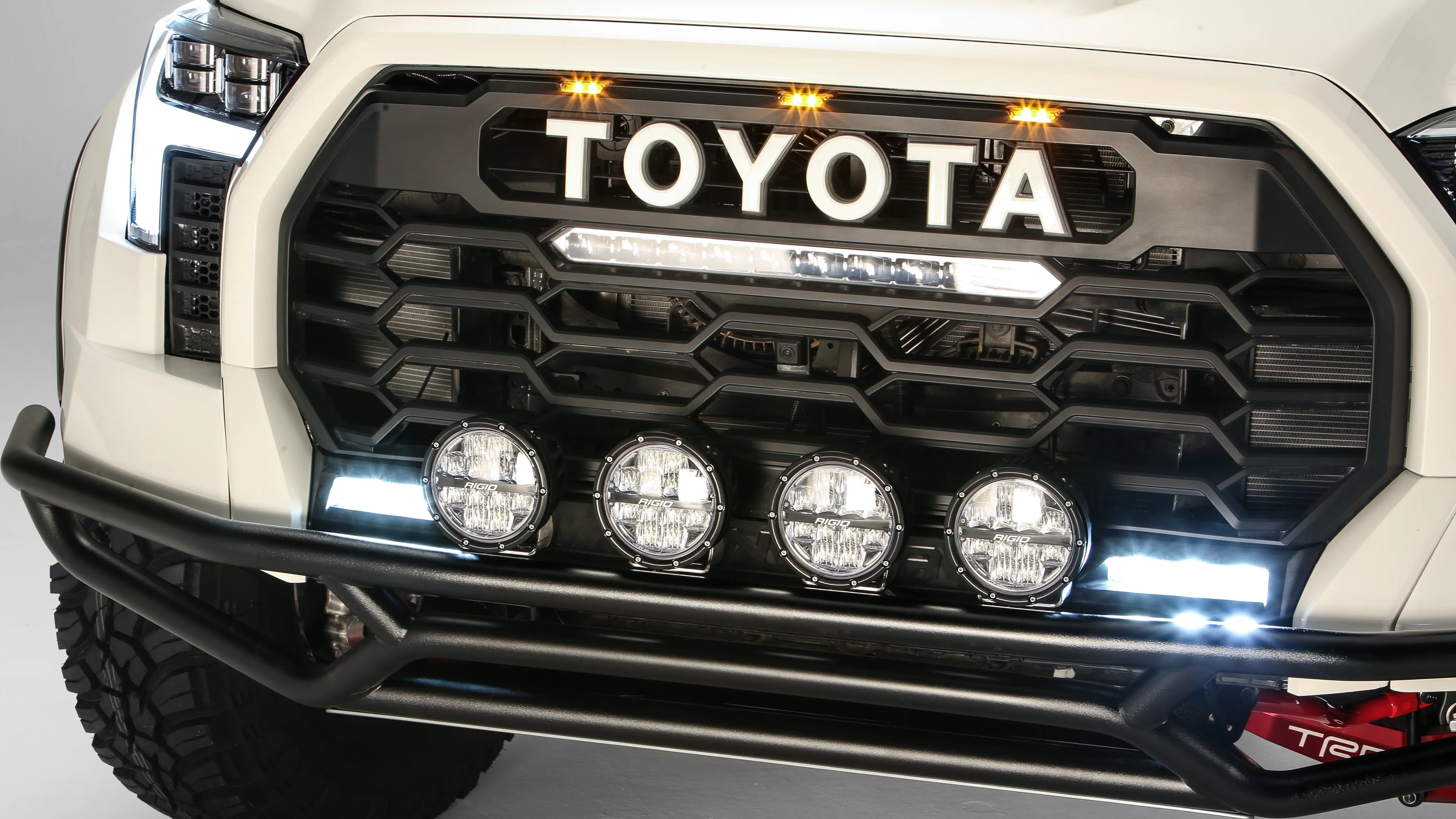 Front grille of a white Toyota truck with black honeycomb pattern, four round Rigid lights, and a black metal bumper guard.