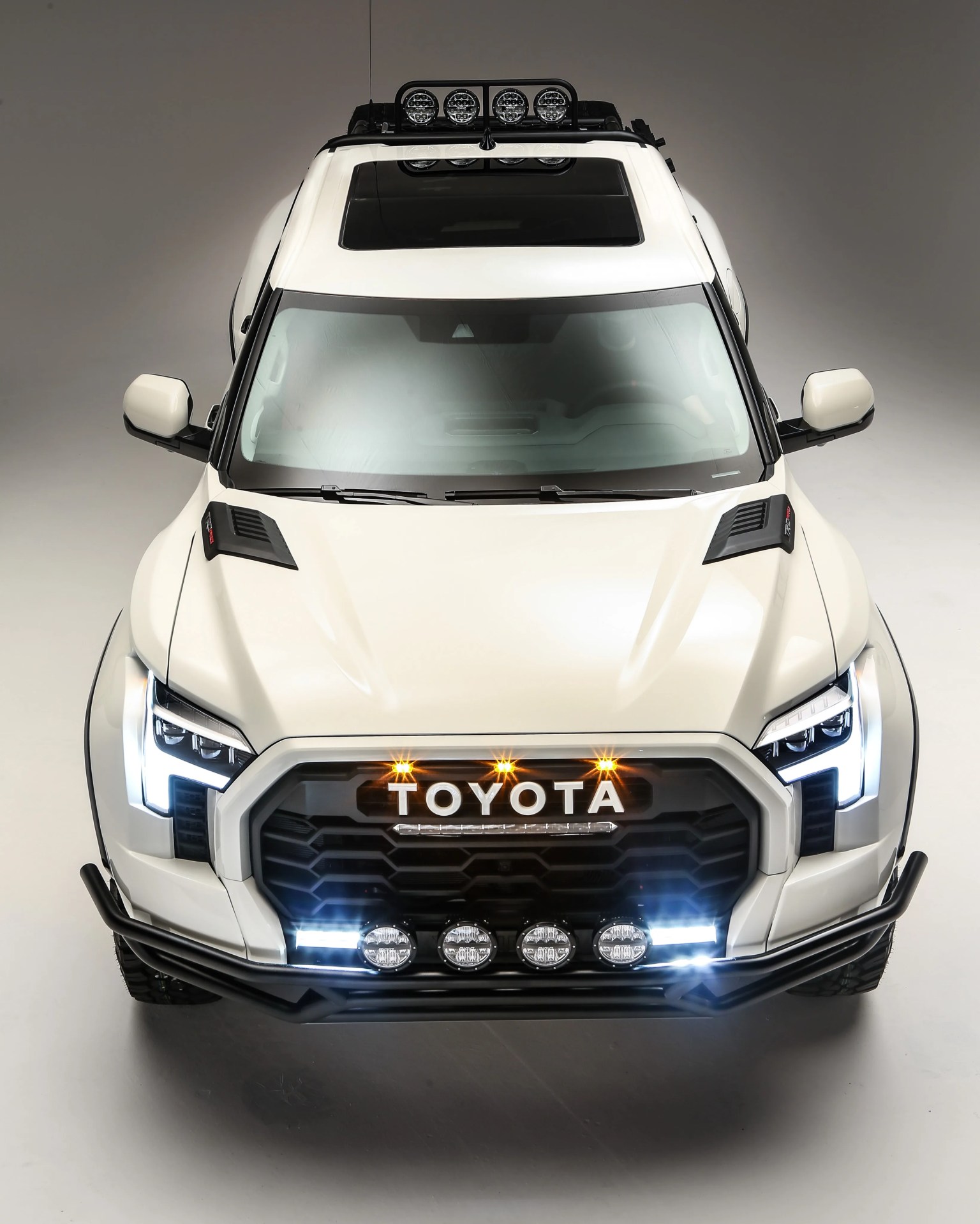White Toyota pickup truck with black grille, roof lights, and off-road bumper in a studio setting.