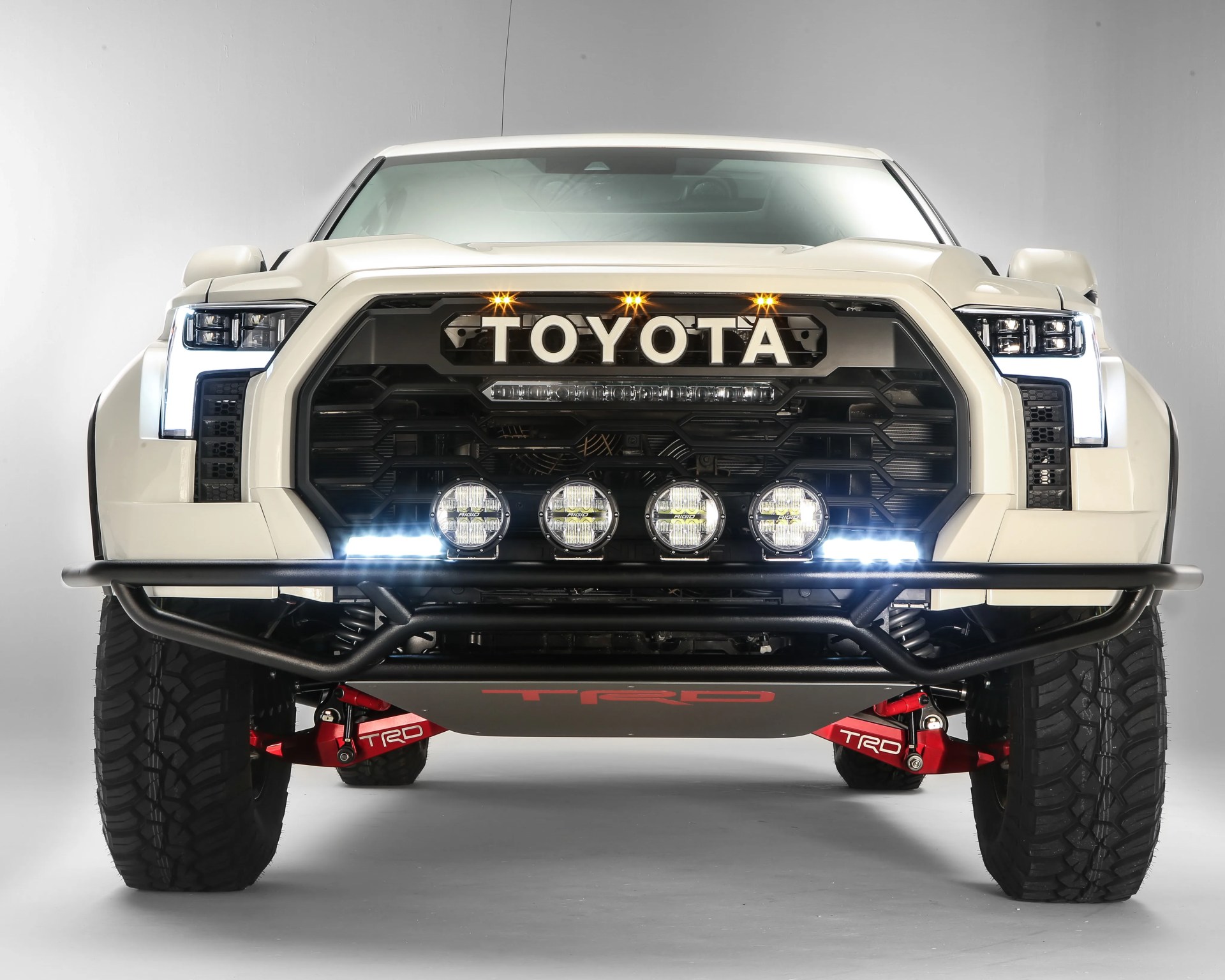 Front view of a white Toyota truck with large off-road tires, black grille, four round auxiliary lights, and red TRD suspension components.