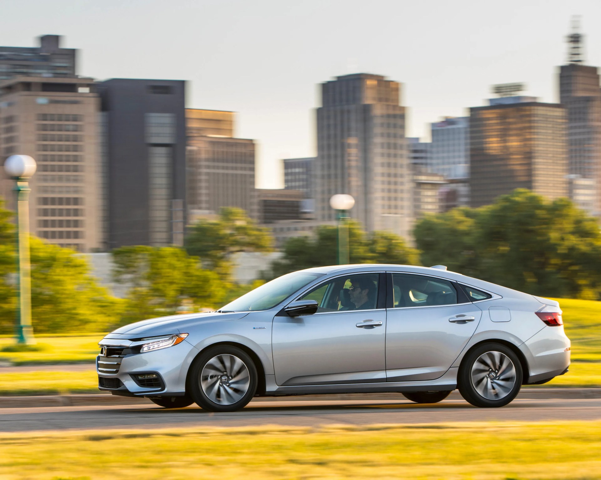 Silver Honda sedan driving on a city street with blurred buildings and greenery in the background.