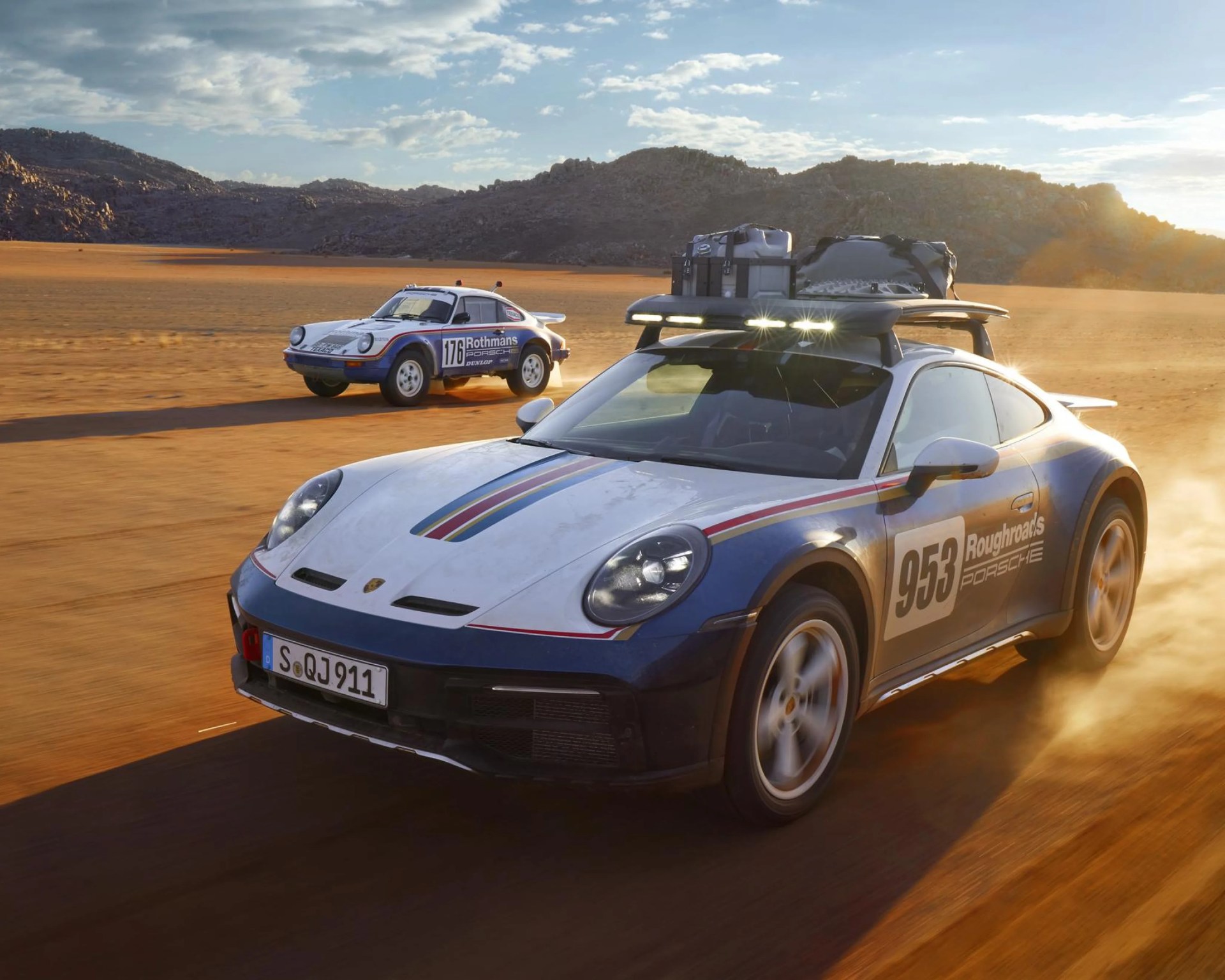 Two Porsche rally cars with vintage racing liveries driving on a dusty desert road, one with roof rack and lights.