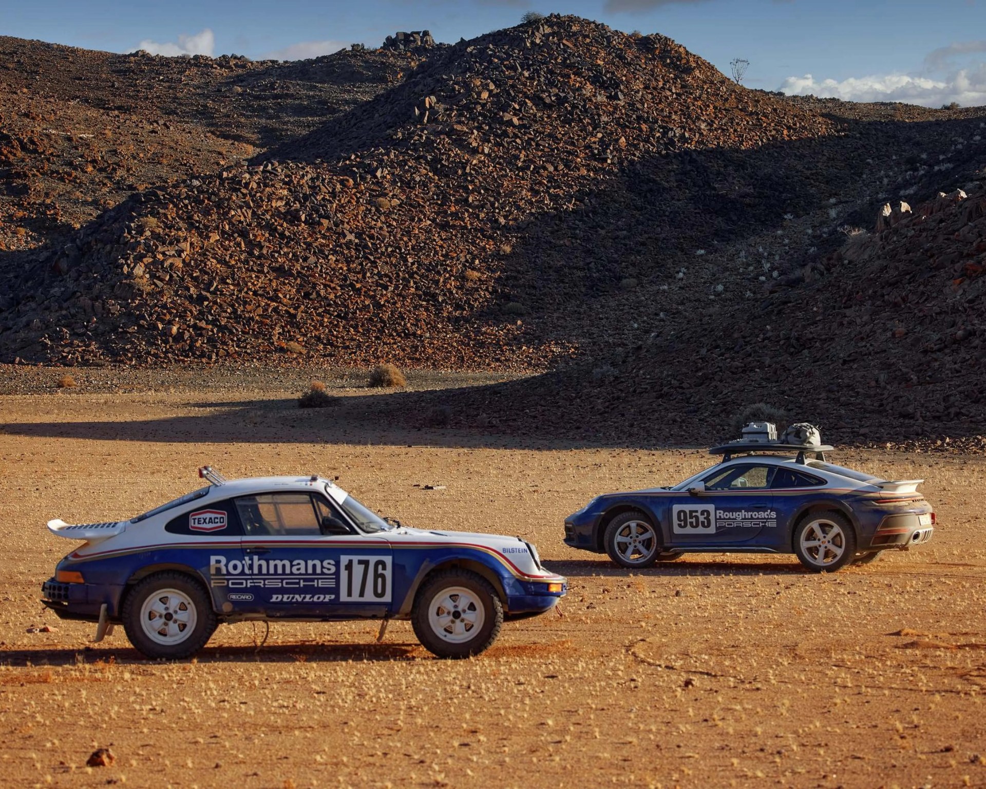 Two blue and white Porsche rally cars with racing decals parked on a rocky desert terrain with hills in the background.