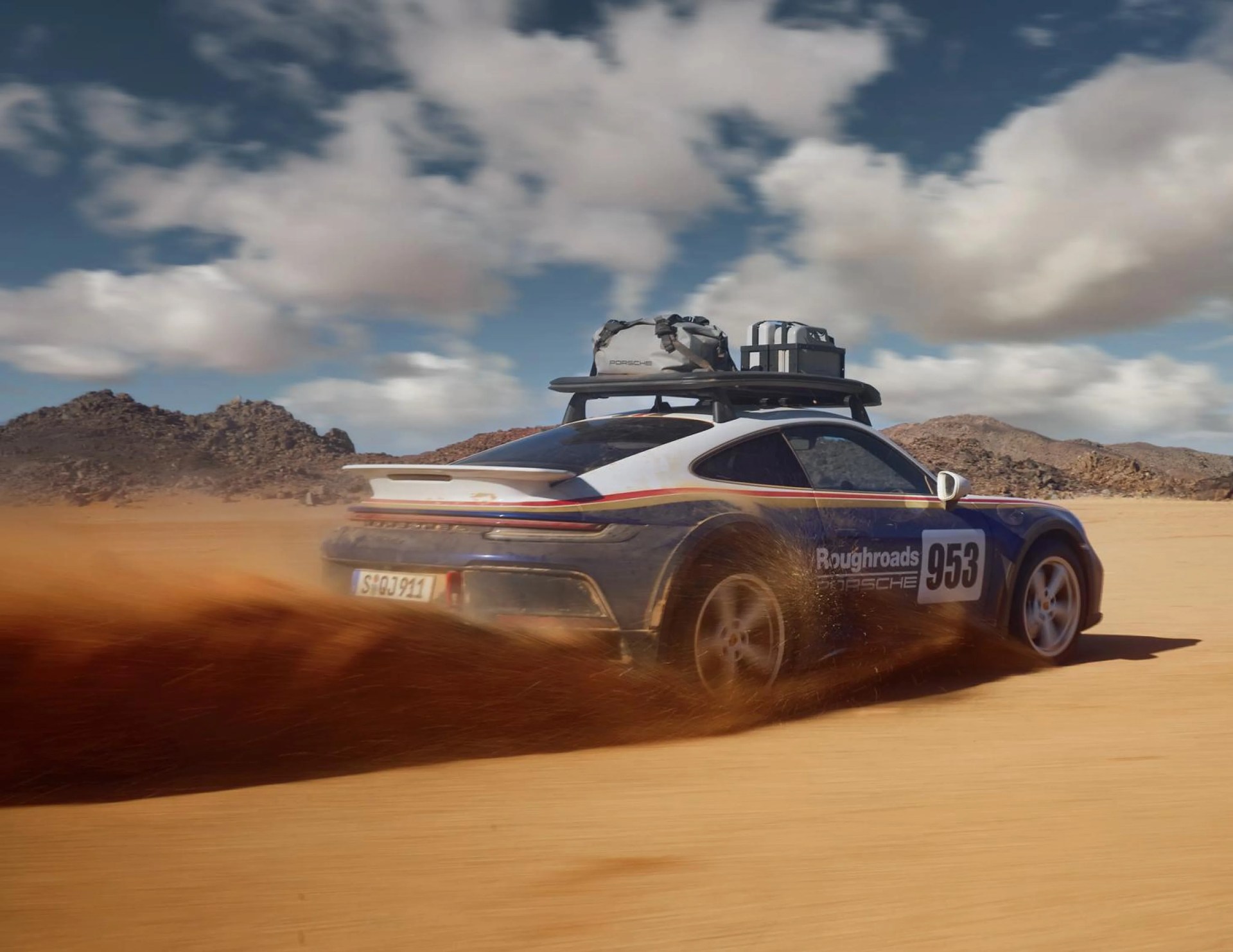Blue and white Porsche 911 with roof rack and gear kicking up dust on a desert dirt road.