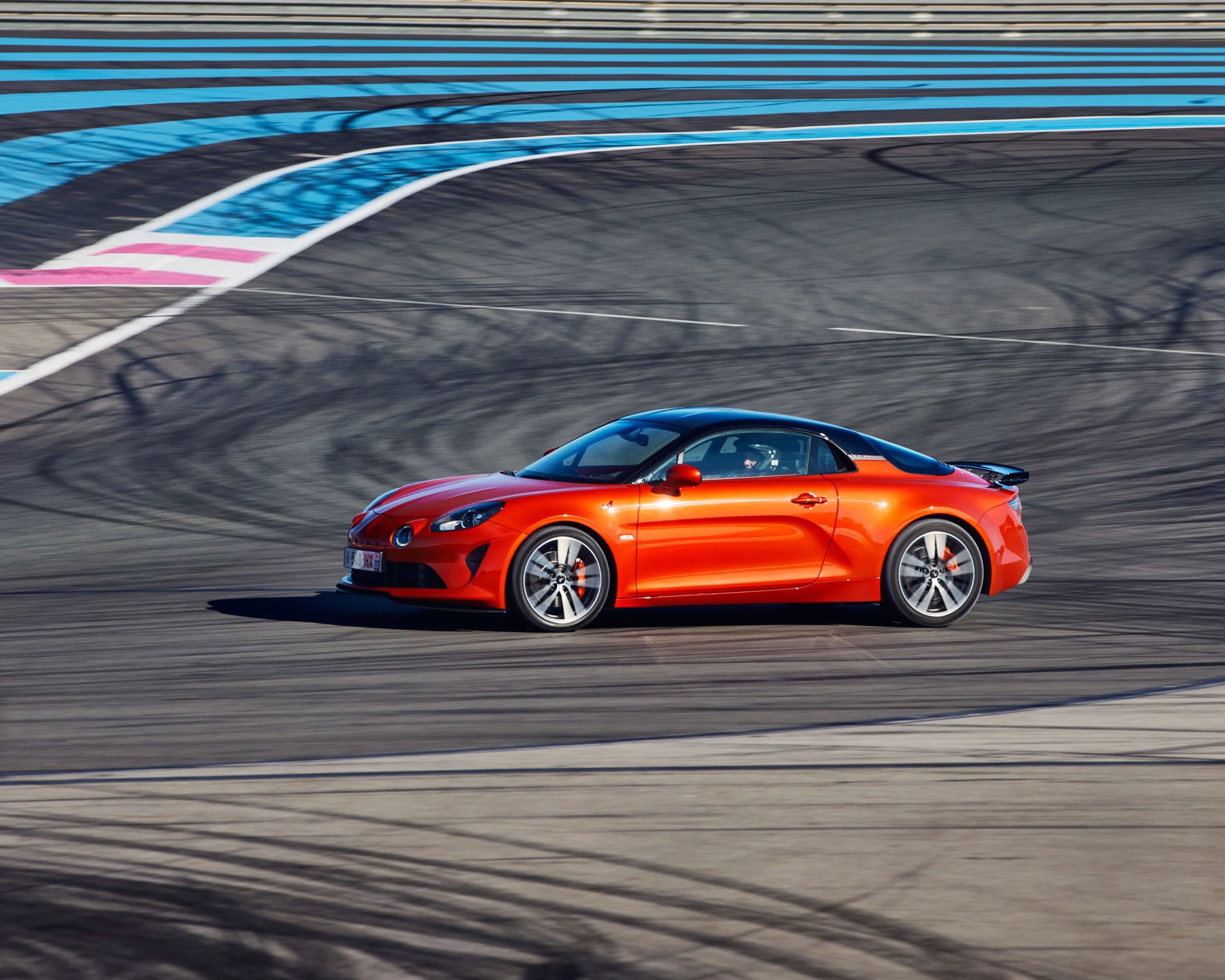 Orange sports car with black roof and silver wheels driving on a racetrack with blue and pink markings.
