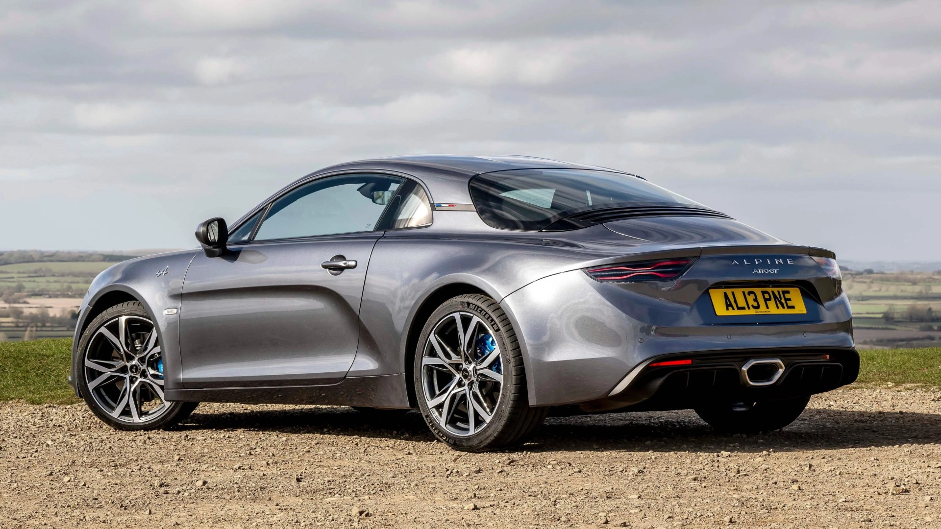 Gray Alpine A110 GT sports car parked on gravel with countryside in the background.