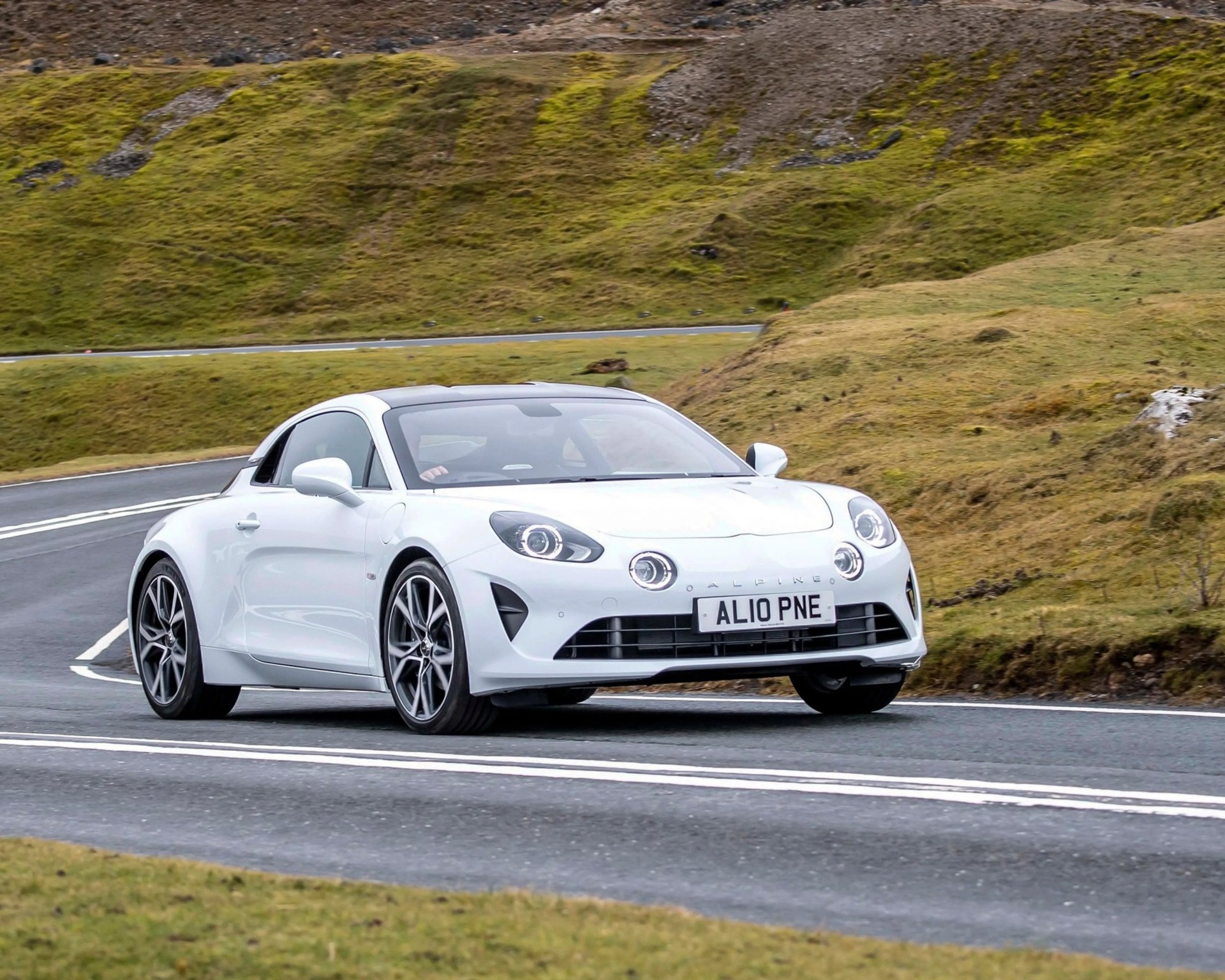 White Alpine sports car with distinctive round headlights driving on a winding road through grassy hills.
