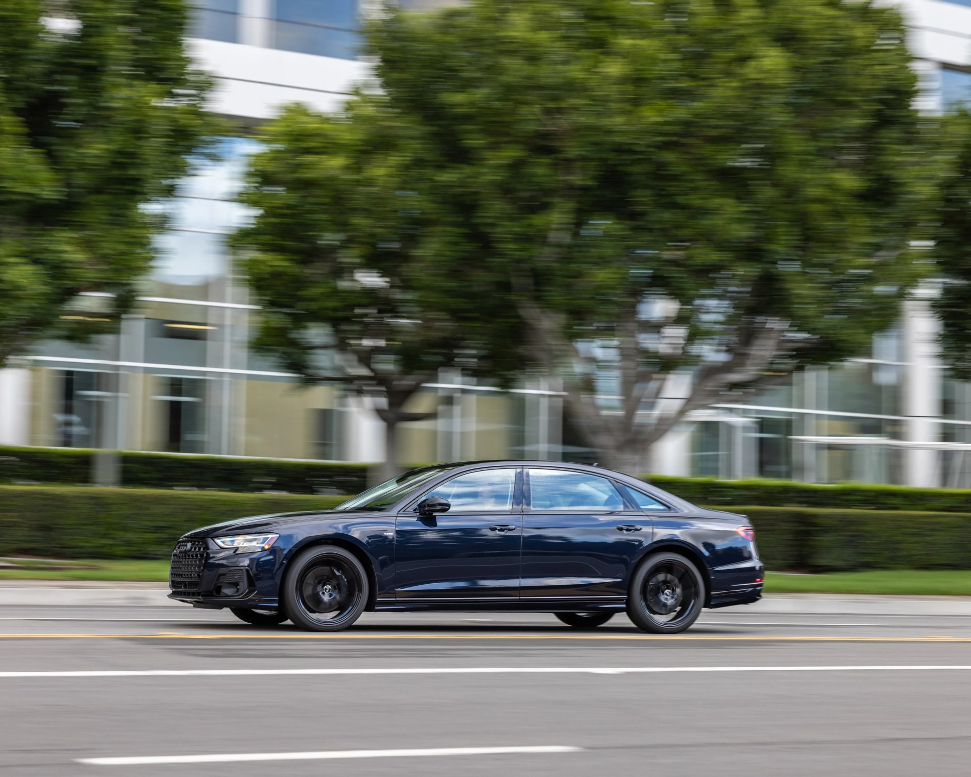 Dark blue luxury sedan driving on a city street with blurred trees and buildings in the background.
