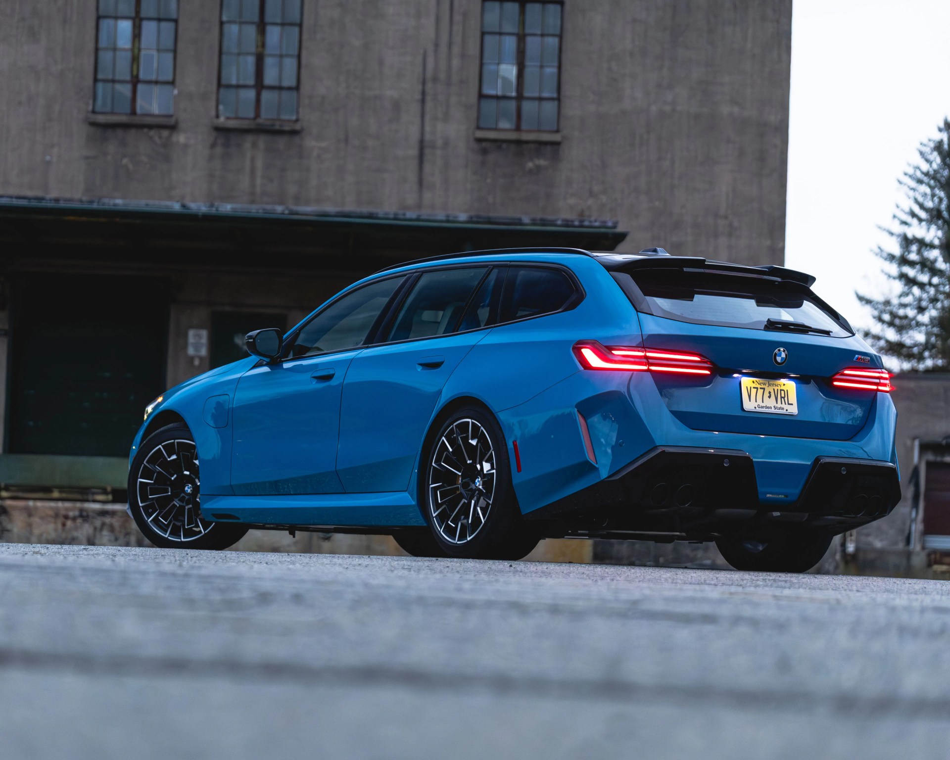 Blue BMW M3 Touring with black wheels and illuminated rear lights parked on a concrete surface near an industrial building.