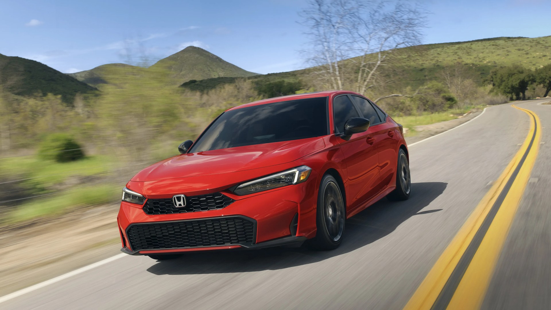 Red Honda sedan with black grille and tinted windows driving on a winding road with green hills in the background.