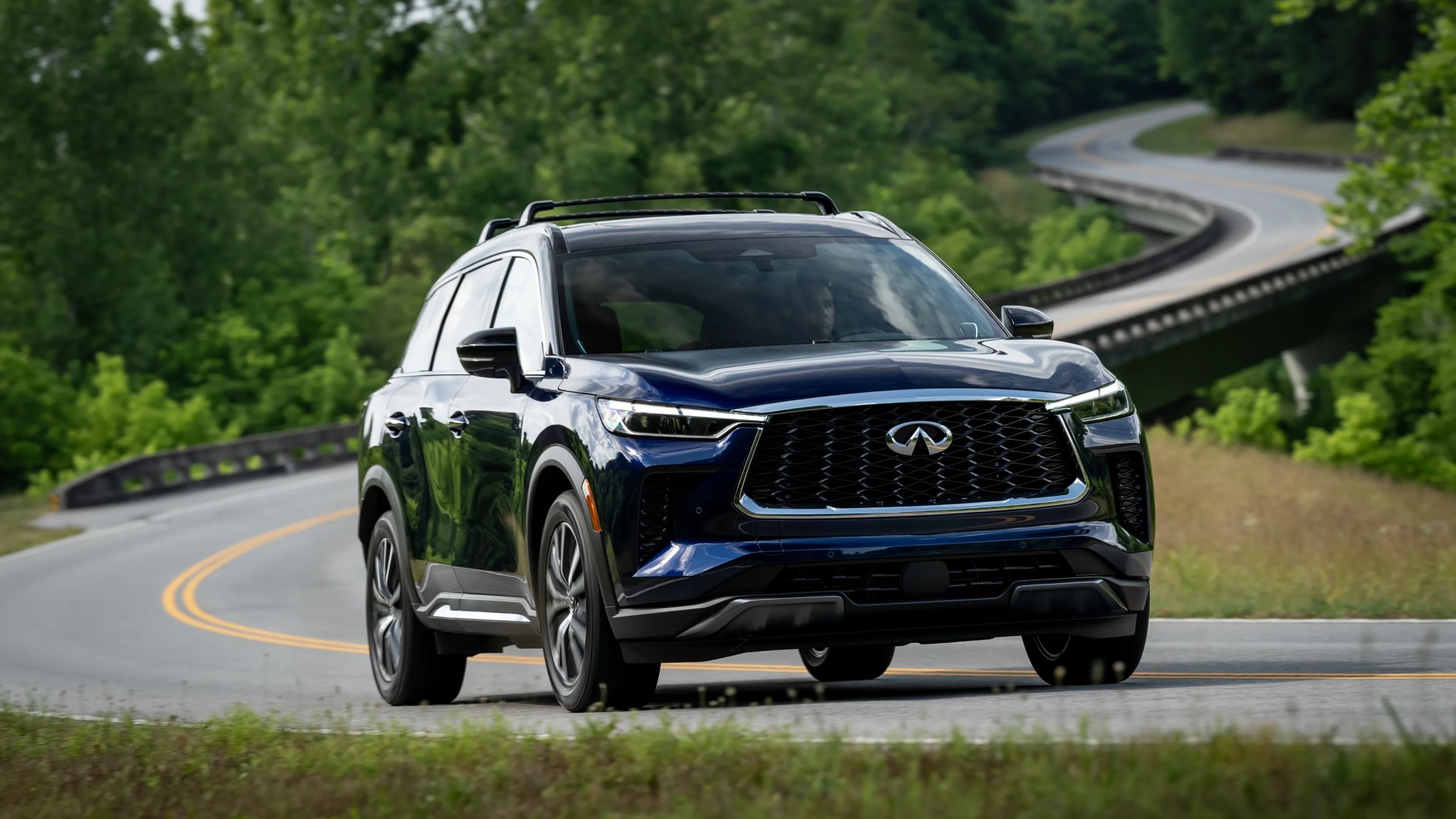 Dark blue Infiniti SUV driving on a winding road surrounded by green trees.