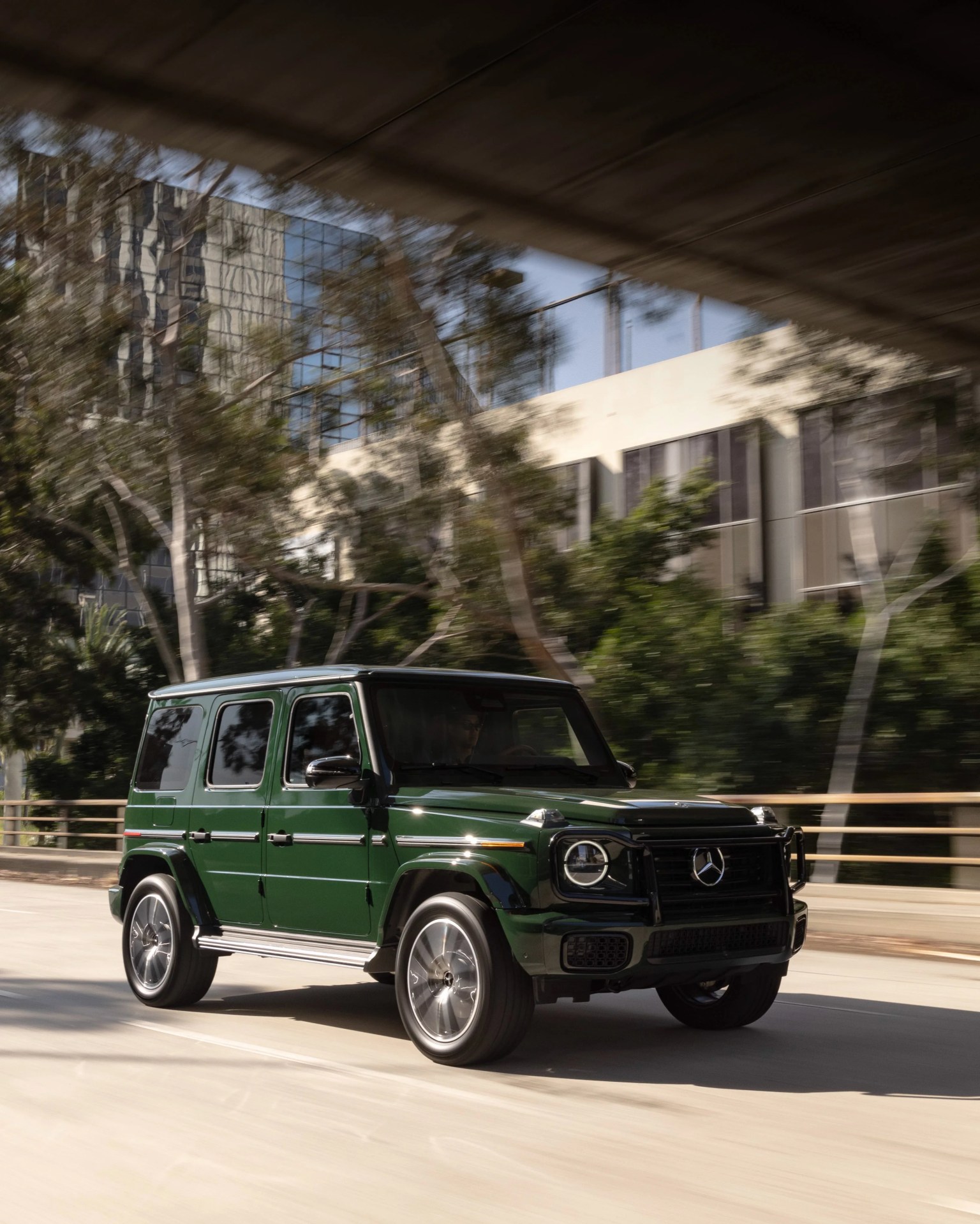 Dark green Mercedes-Benz G-Class SUV driving on a city street under an overpass.