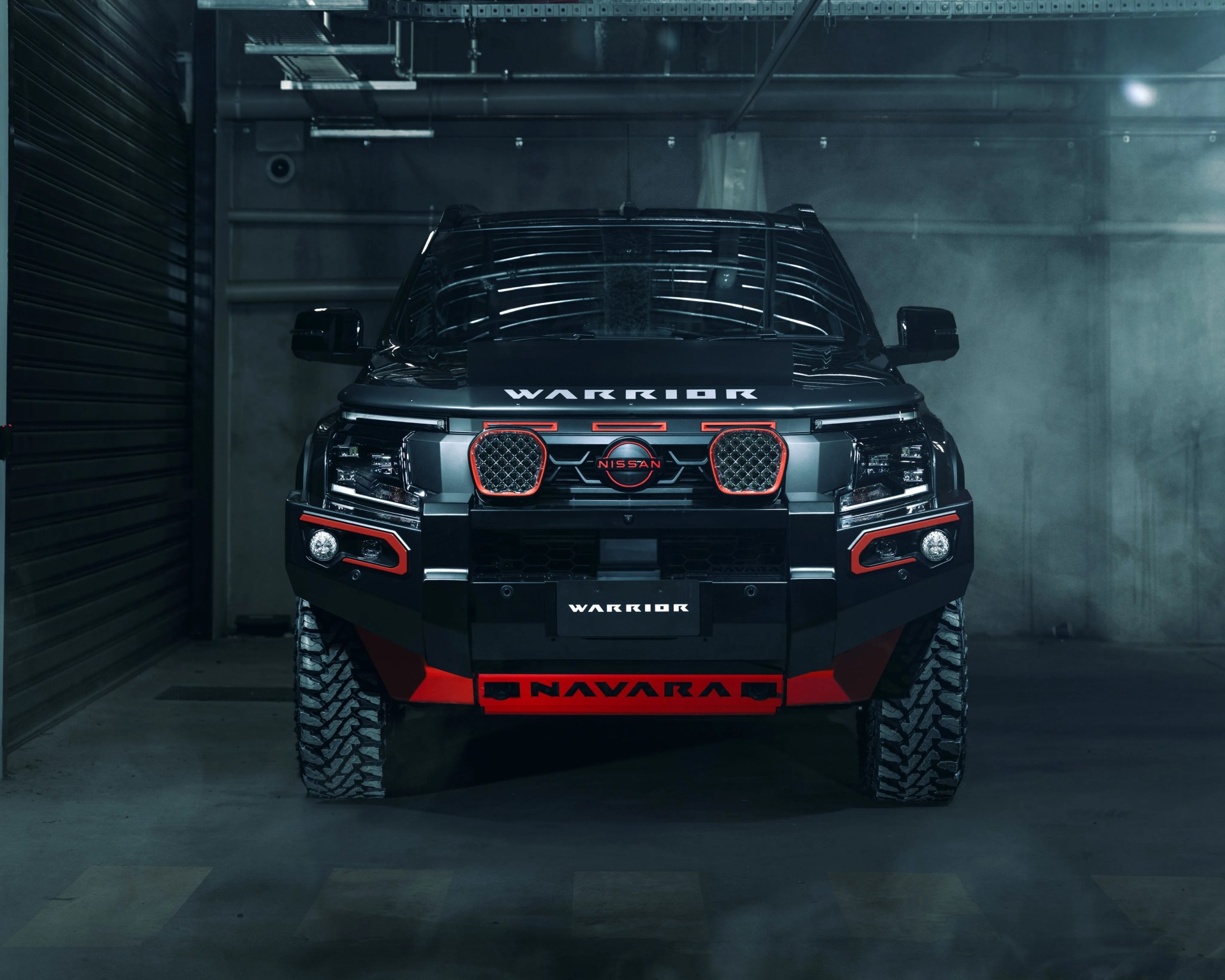 Front view of a black Nissan Navara Warrior pickup truck with red accents and rugged tires in a dimly lit garage.