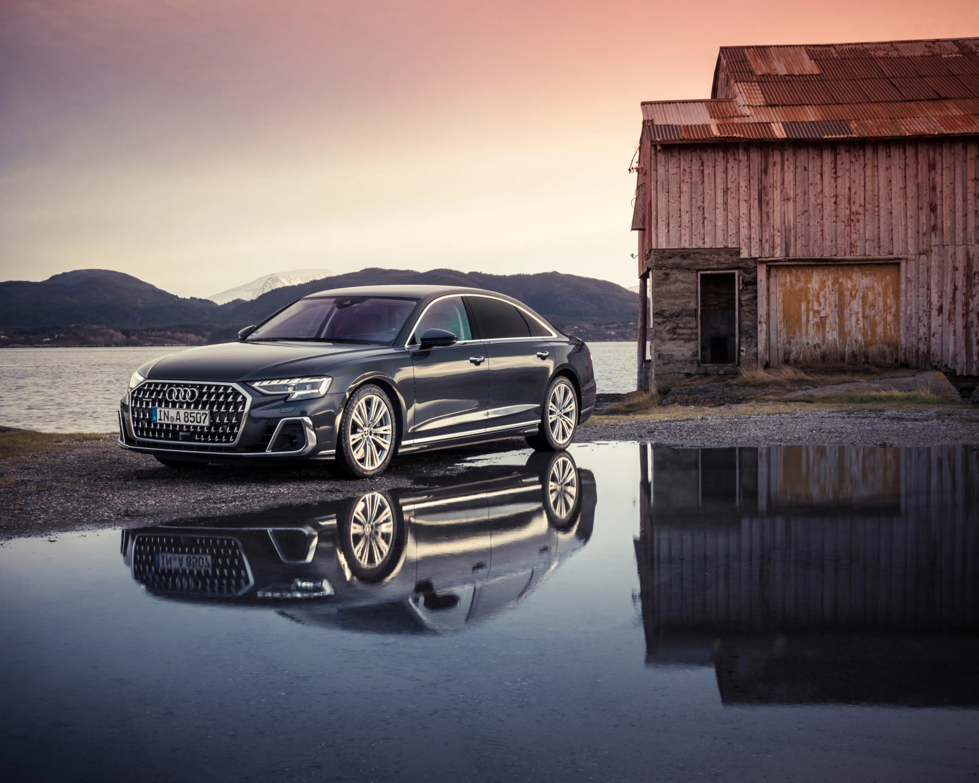 Black Audi sedan parked near a weathered wooden building with its reflection in a puddle at sunset.