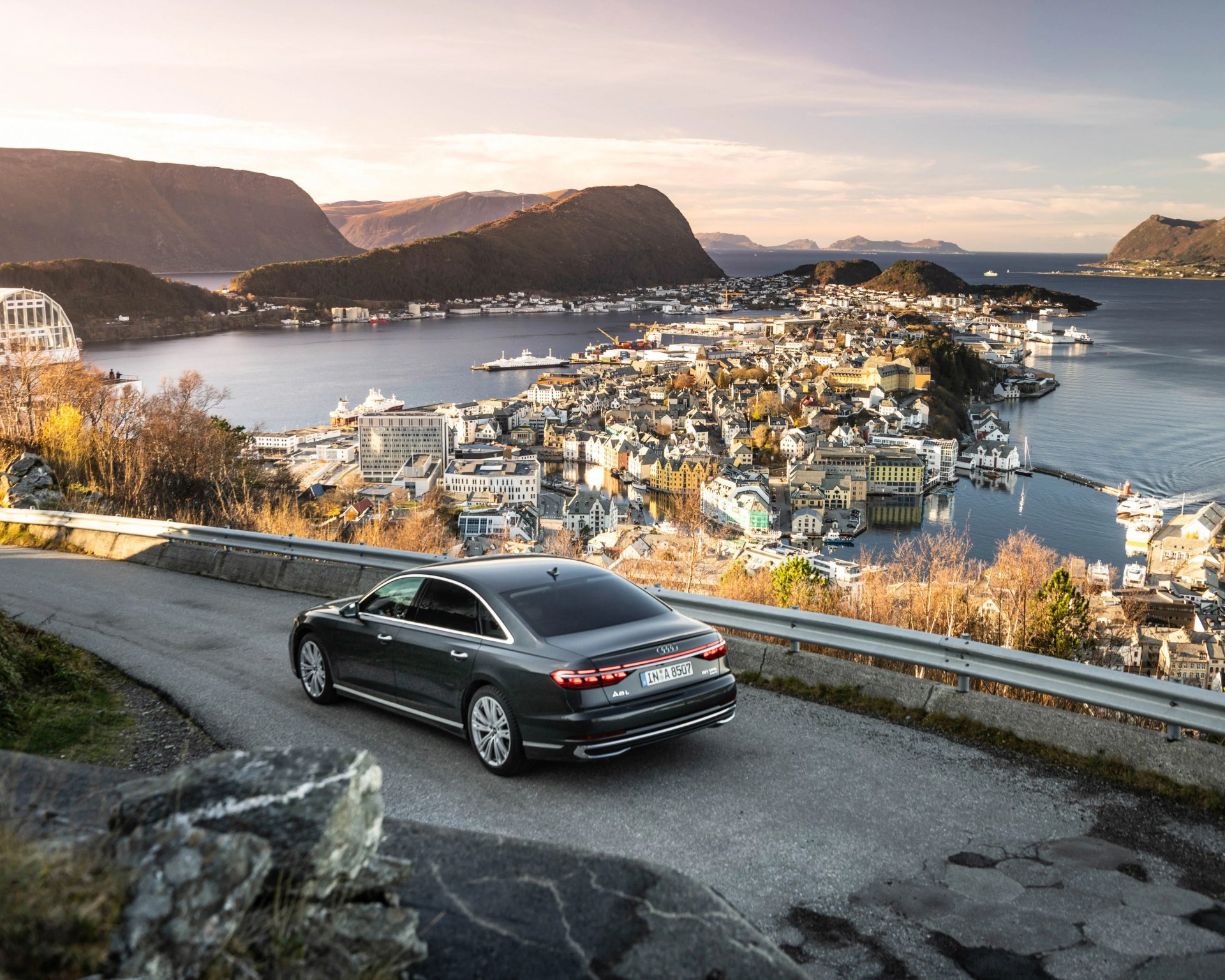 Dark gray Audi A8L sedan driving on a mountain road overlooking a coastal town and fjord.
