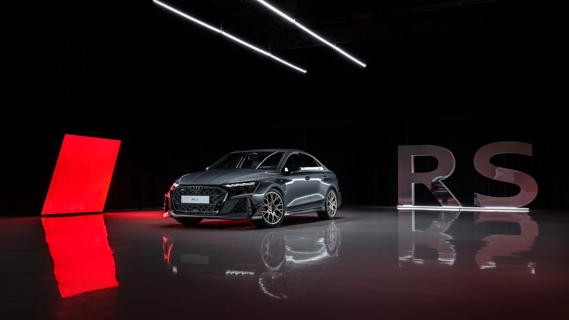 Dark gray Audi RS3 sedan with intricate alloy wheels in a dimly lit studio with red and white light accents.