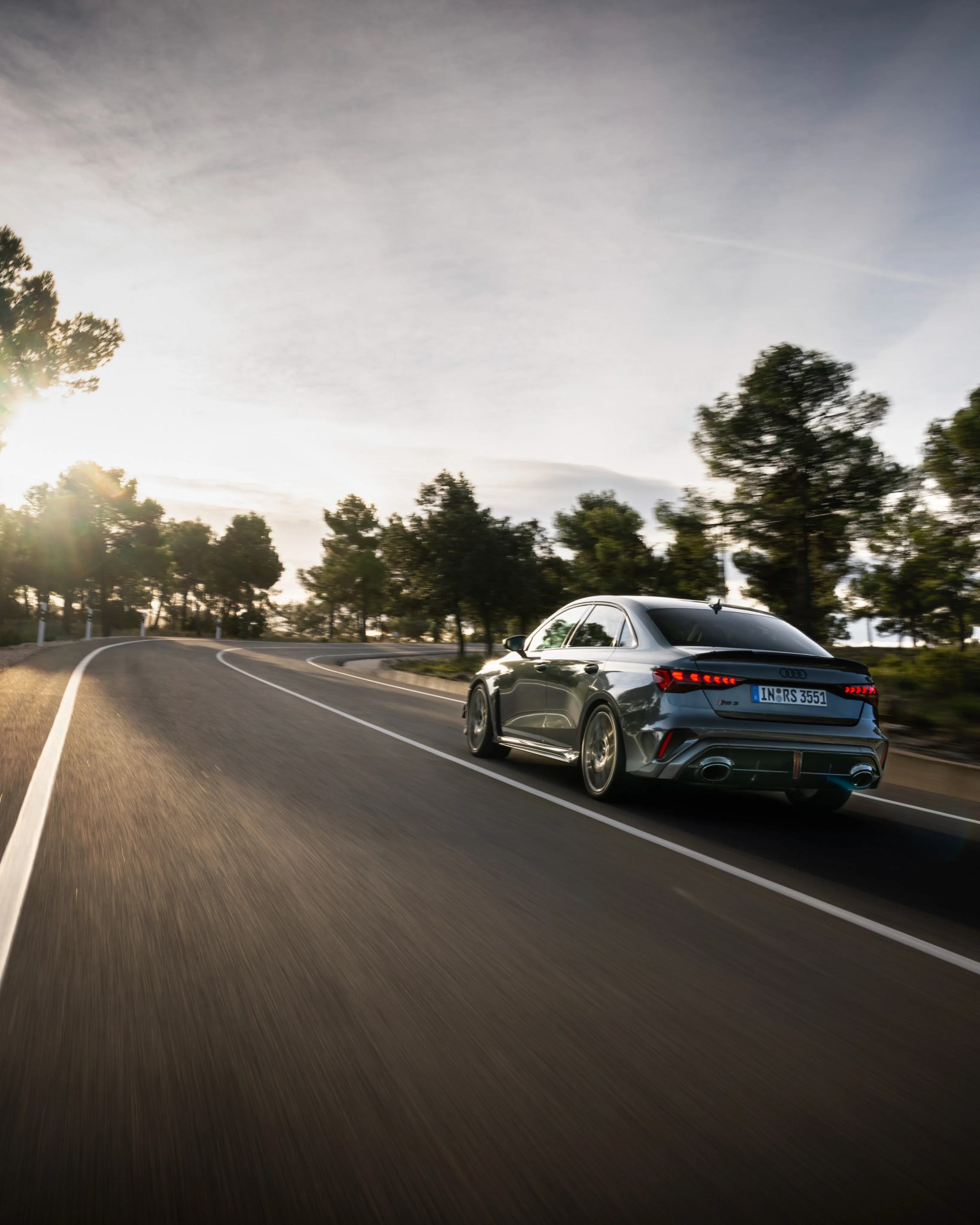 Gray Audi RS3 sedan driving on a winding road with trees and sunlight in the background.